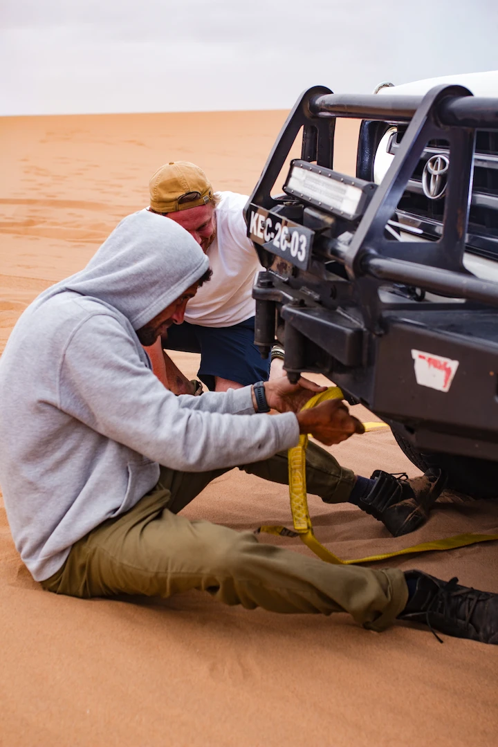 Twee mensen zijn samen aan het werk in de woestijn en maken een gele sleepkabel vast aan de voorkant van een witte terreinwagen. De ene draagt een grijze hoodie en de andere een beige pet en een wit shirt. Ze worden omringd door zand.