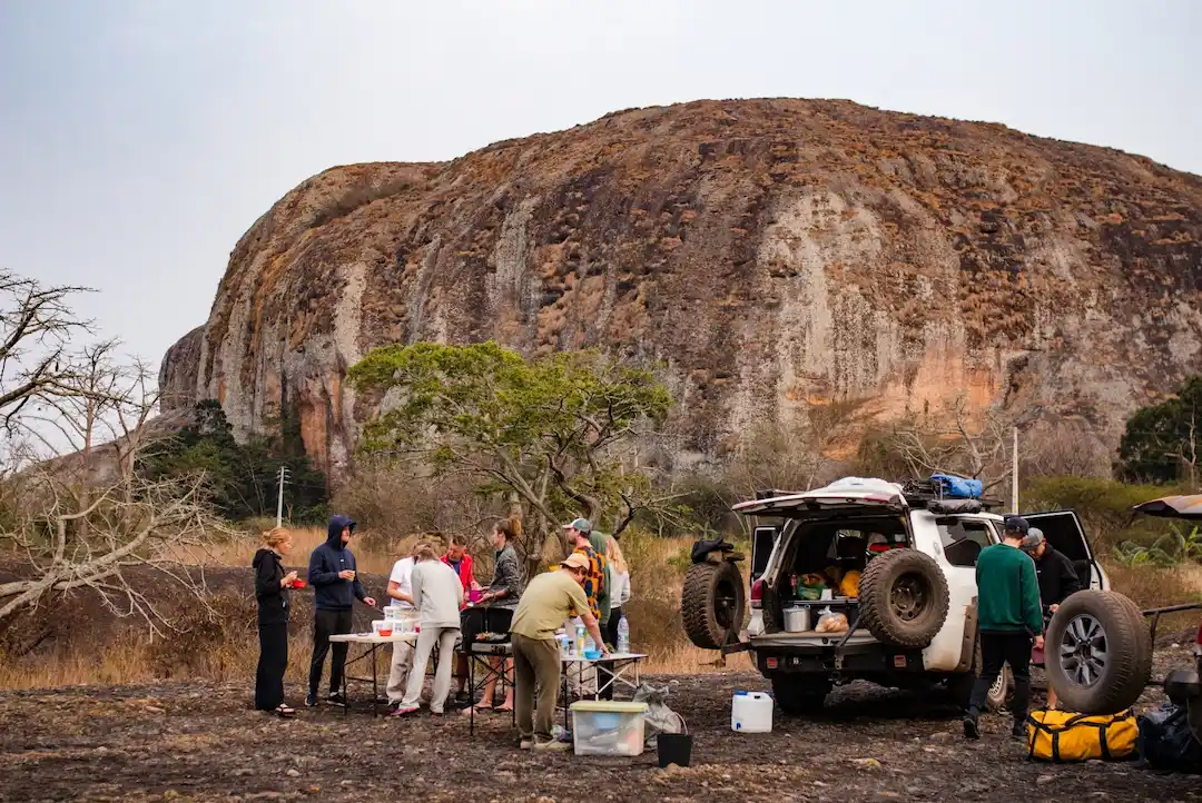 Een groep mensen staat bij tafels en een terreinwagen en maakt eten klaar op een kampeerterrein. Op de achtergrond rijst een grote rotsachtige heuvel op onder een bewolkte hemel. Overal liggen kampeerspullen en proviand verspreid.