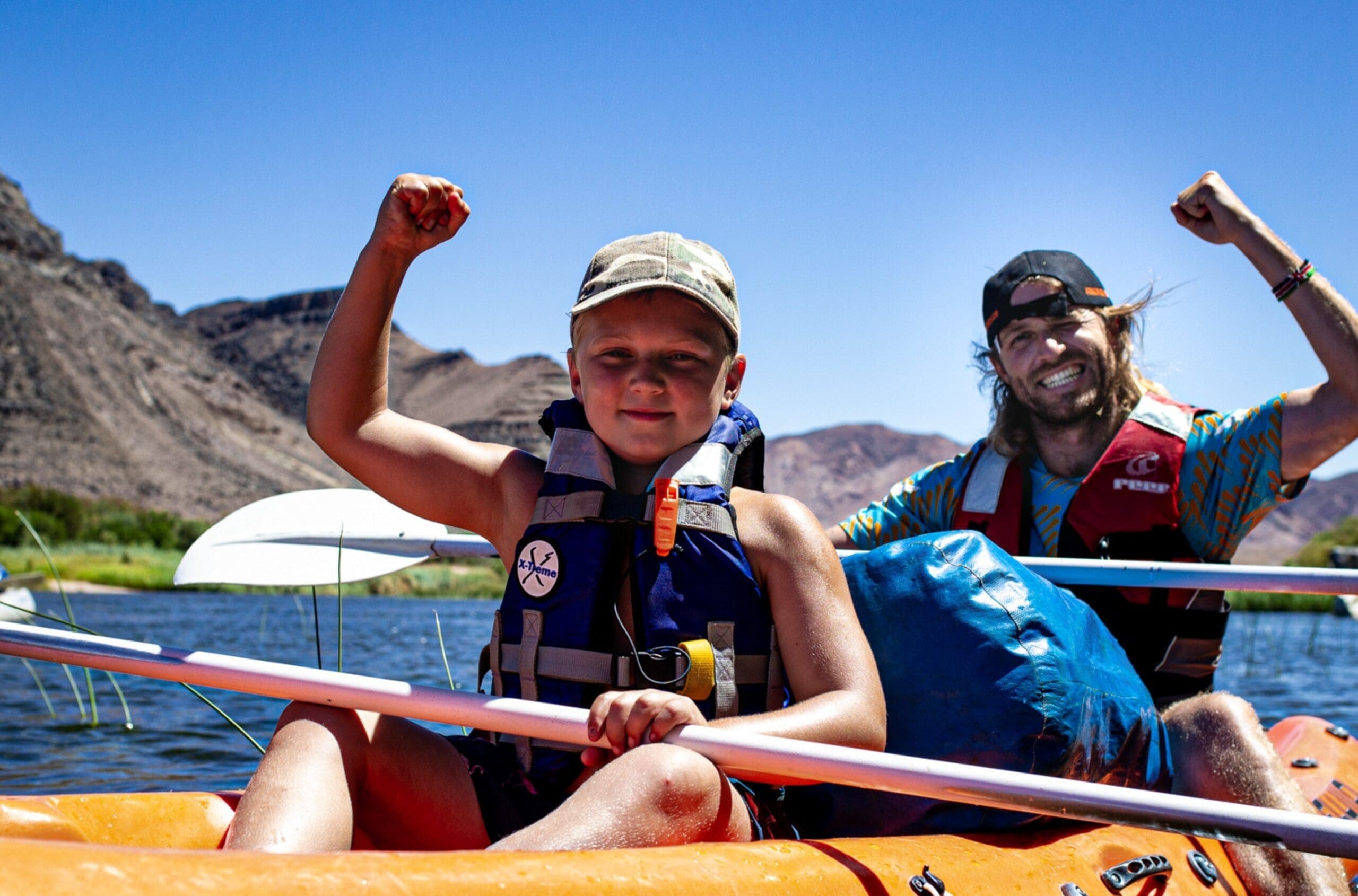 A smiling man and a boy, both wearing life jackets, paddle a kayak on a lake with mountains in the background. They raise their arms in a gesture of excitement under a clear blue sky.