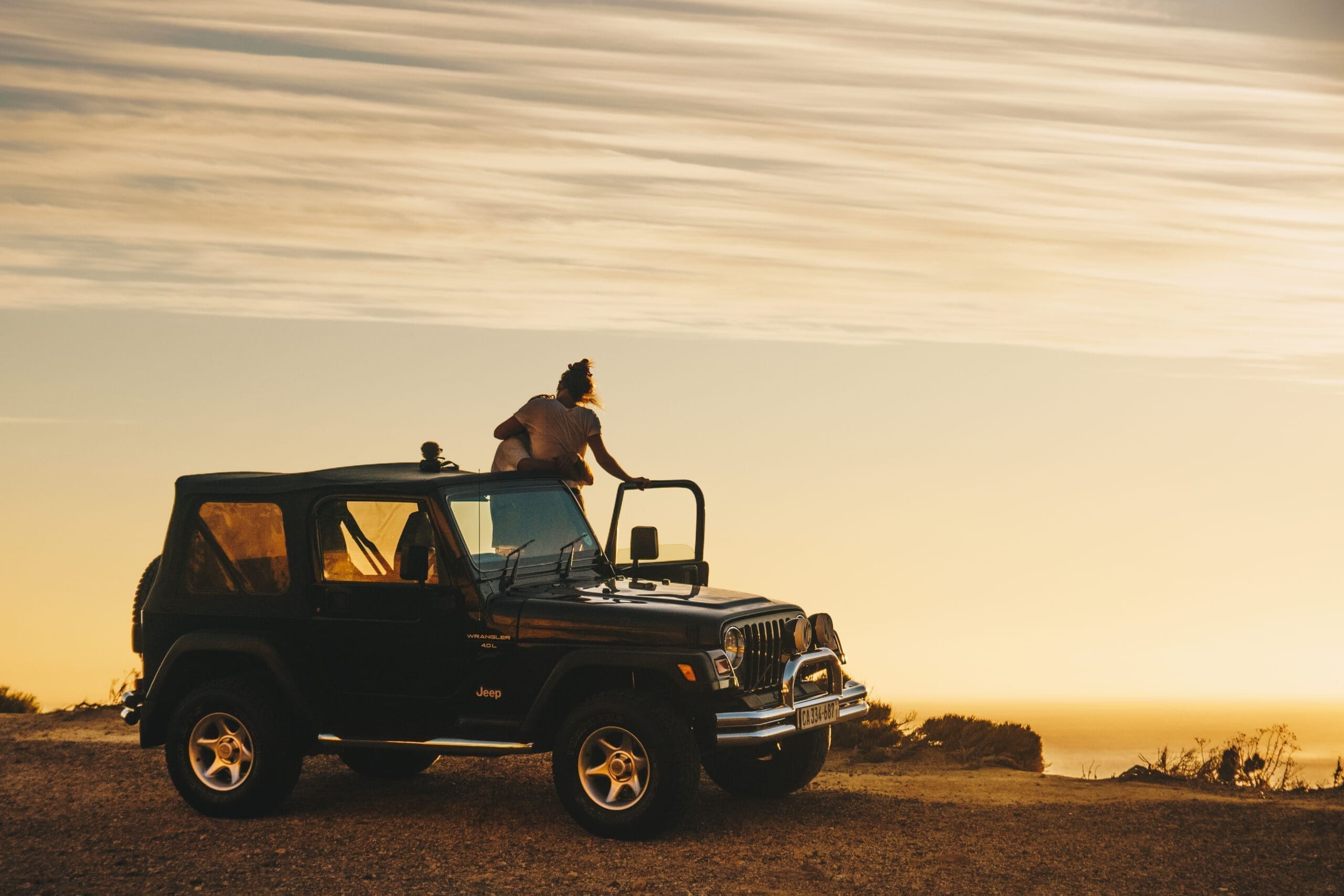 Een persoon zit op het dakframe van een zwarte Jeep die geparkeerd staat op een zandweg, kijkend naar een gouden zonsondergang met gestreepte wolken vlakbij de kust - perfect om te reizen in Zuid Afrika.