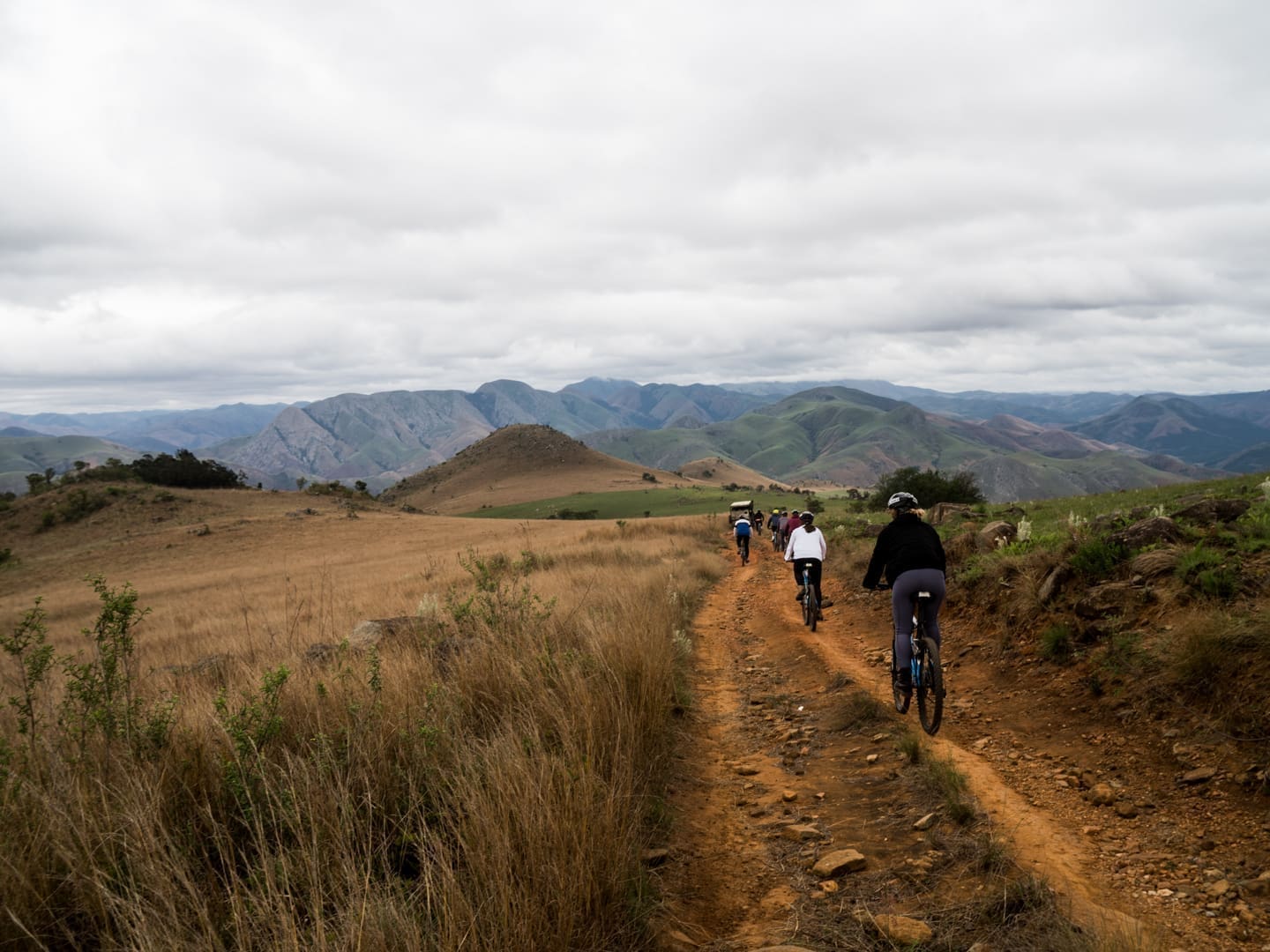 een groep fietsers rijdt over een stoffig pad door een heuvelachtig landschap in eswatini omringd door bergen onder een bewolkte lucht