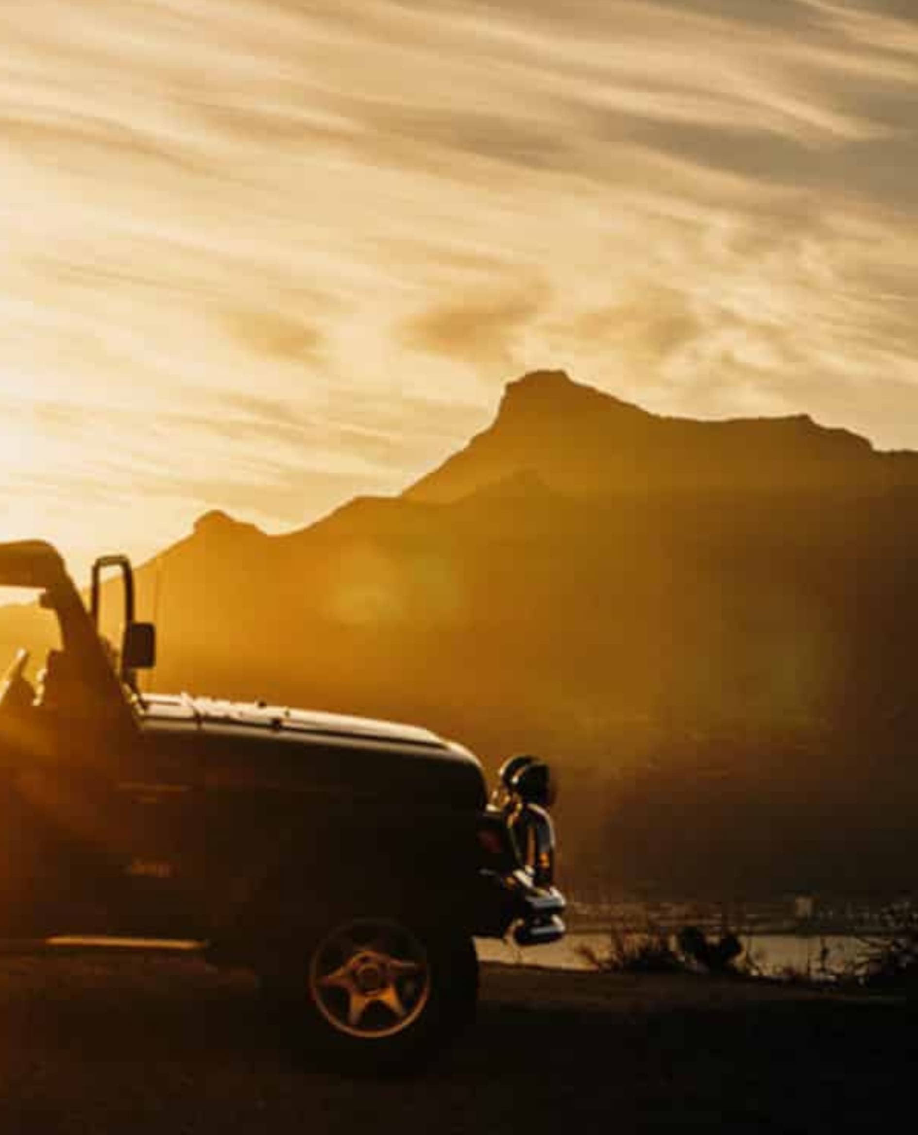 A black SUV is parked with its front facing left at sunset, with mountains silhouetted in the background and a golden sky overhead.