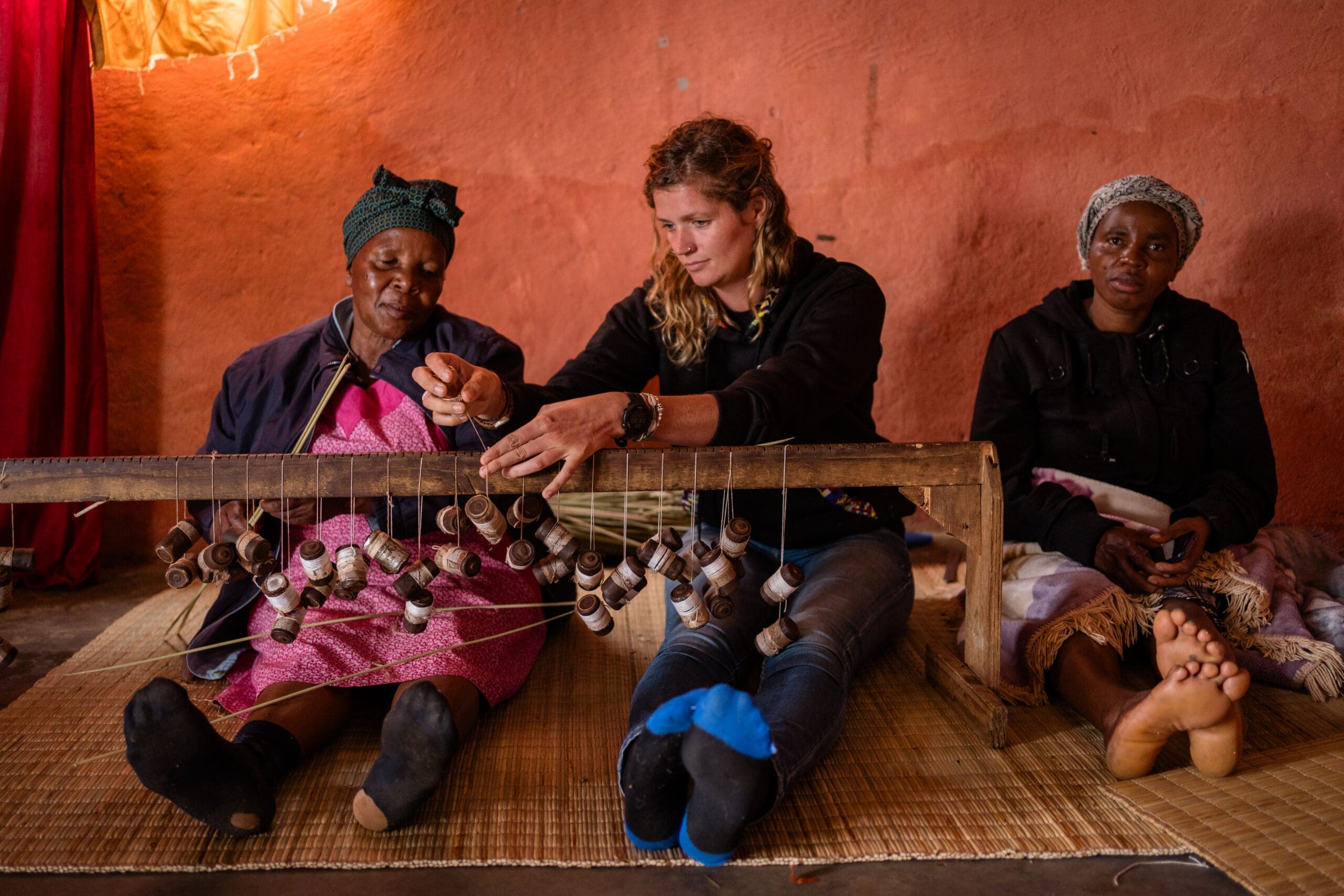 Drie vrouwen zitten binnenshuis op matten in eSwatini; twee werken samen aan een traditioneel weefgetouw met houten spoelen, terwijl de derde vrouw toekijkt. Deze scène biedt reizigers een authentieke blik op lokaal vakmanschap tegen een aardse, oranje muur.