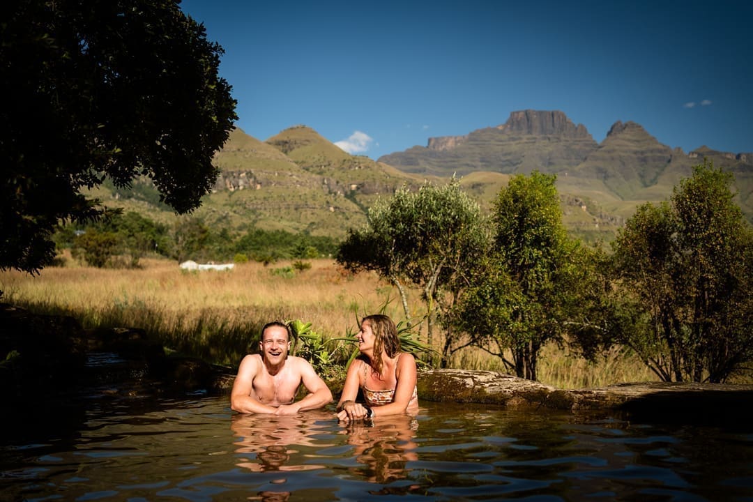 Een man en een vrouw ontspannen en glimlachen in een natuurlijk zwembad, omringd door groen en bergen onder een helderblauwe hemel. Deze vredige scène toont reizigers die genieten van de schoonheid van Zuid-Afrika, met majestueuze bergen op de achtergrond.