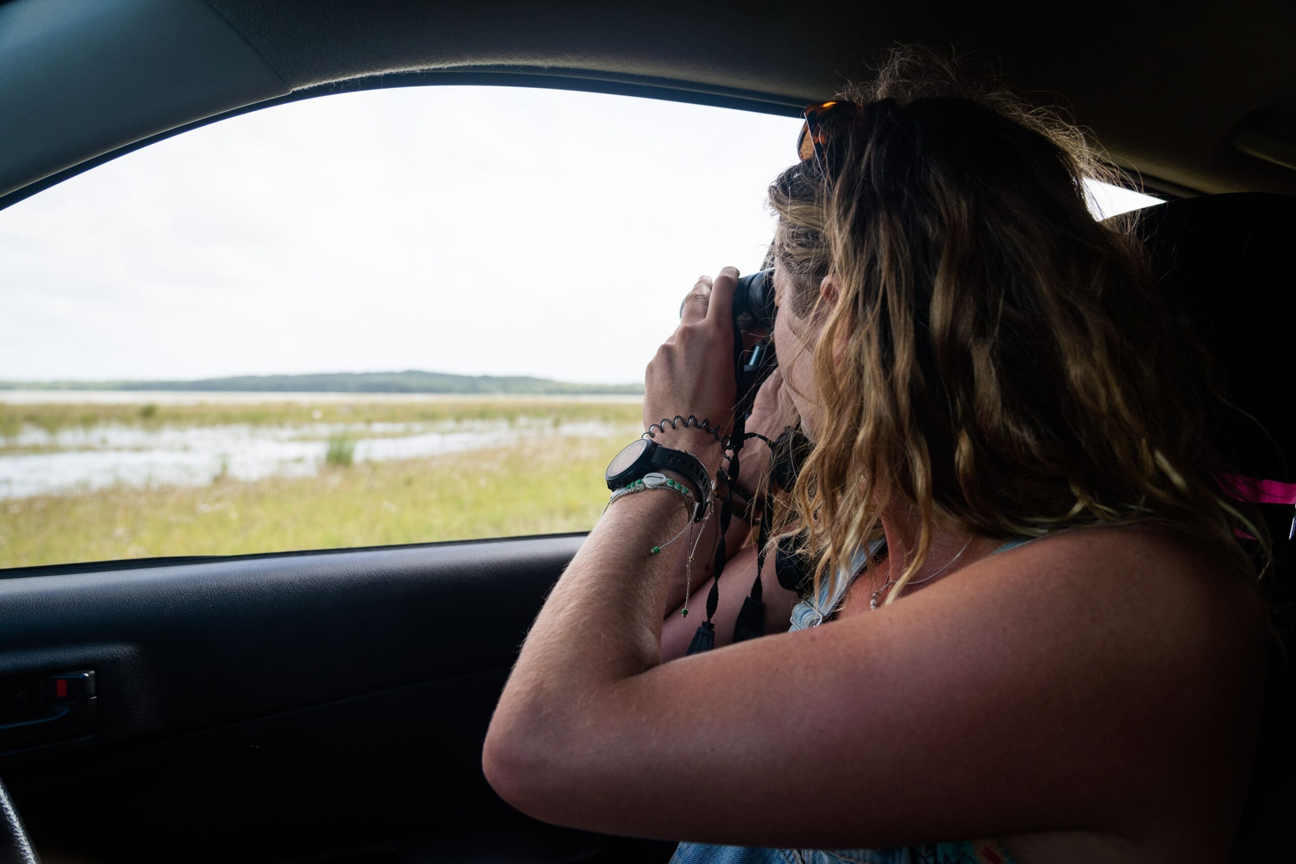 Een persoon met golvend haar zit in een auto in Mozambique, houdt een verrekijker vast en kijkt uit het open raam naar een graslandschap. Hun gezicht is weggedraaid van de camera, waarmee ze de avontuurlijke geest van Afrika vastleggen.