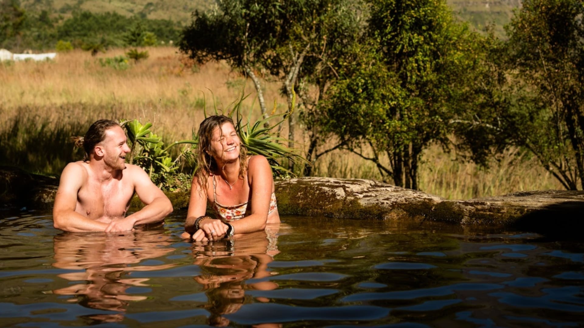 Een man en vrouw ontspannen en glimlachen samen in een natuurlijk buitenzwembad omringd door groen, genietend van het zonlicht als reizigers in Zuid Afrika met bomen die weerspiegeld worden in het glinsterende water.