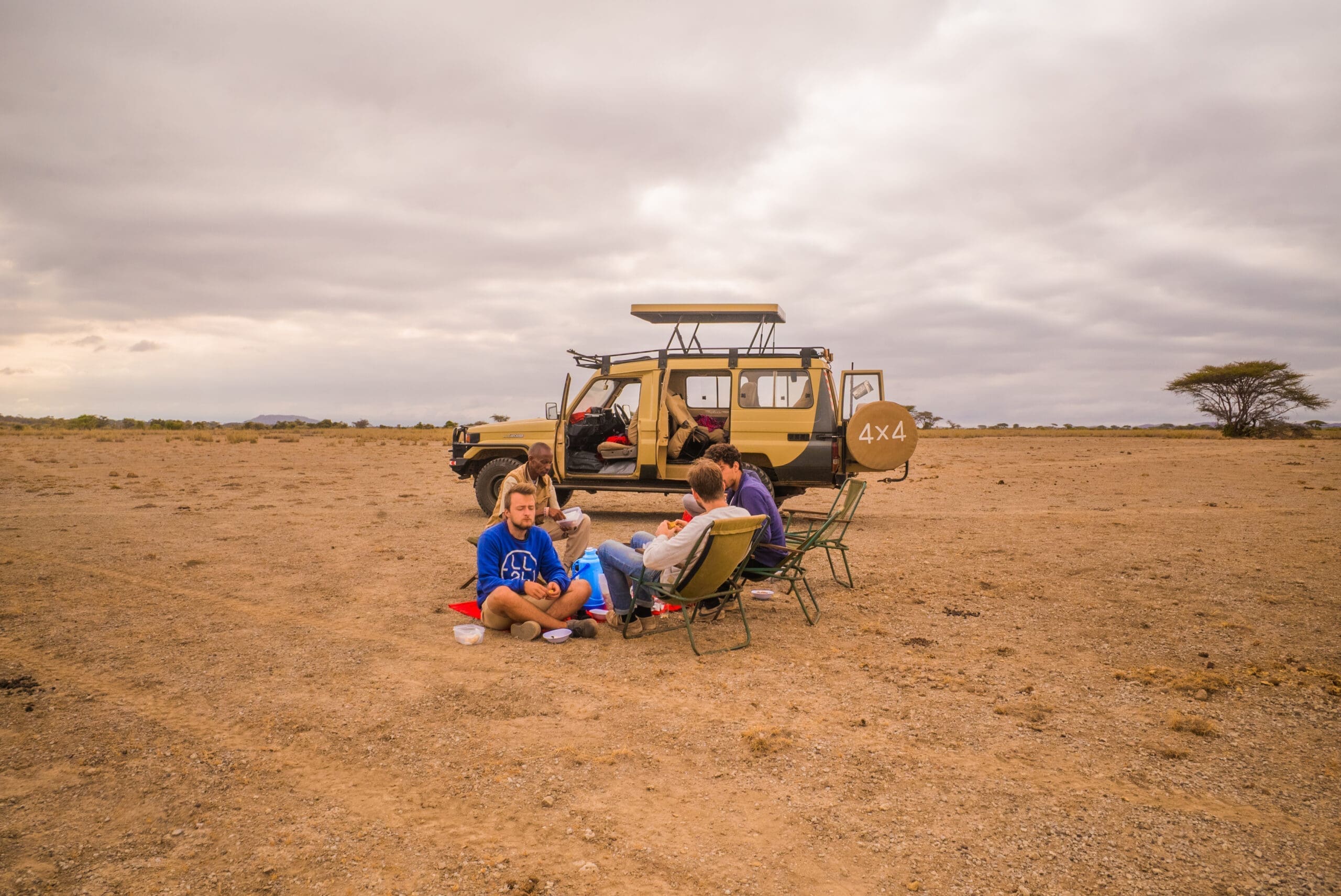 Vier reizigers ontspannen op campingstoelen en op de grond, genietend van een maaltijd naast hun 4x4 safarivoertuig op de uitgestrekte, open vlaktes van Afrika, met een eenzame boom in de verte onder een bewolkte hemel.