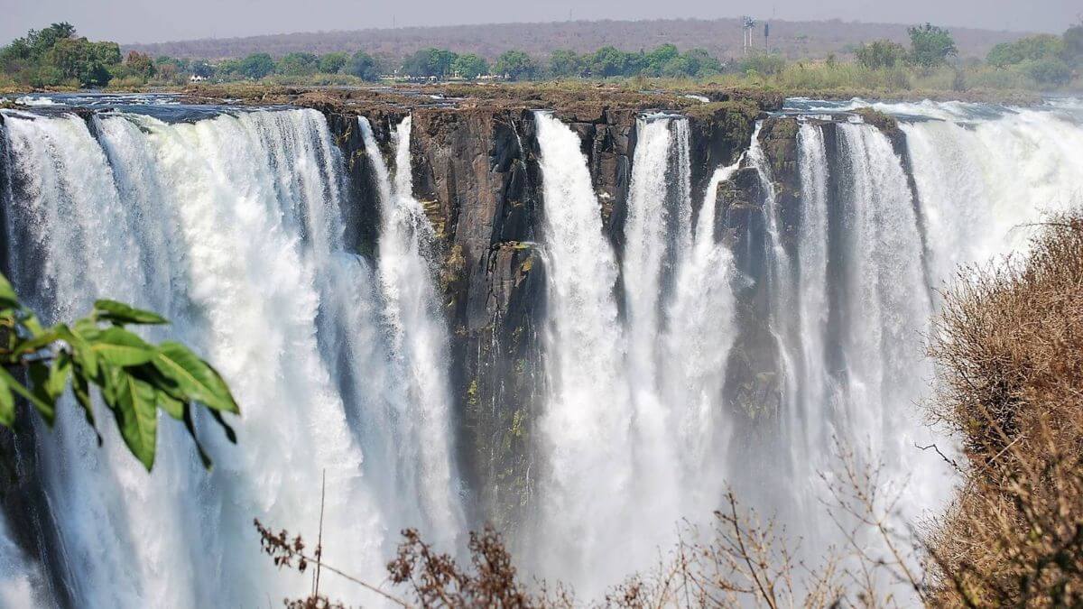Weids uitzicht op de Victoria watervallen op de grens met Botswana, met krachtige watervallen die over een rotsachtige klif klateren, omgeven door groene en bruine vegetatie en mist die opstijgt uit het vallende water - een van Afrika's grootste natuurwonderen.