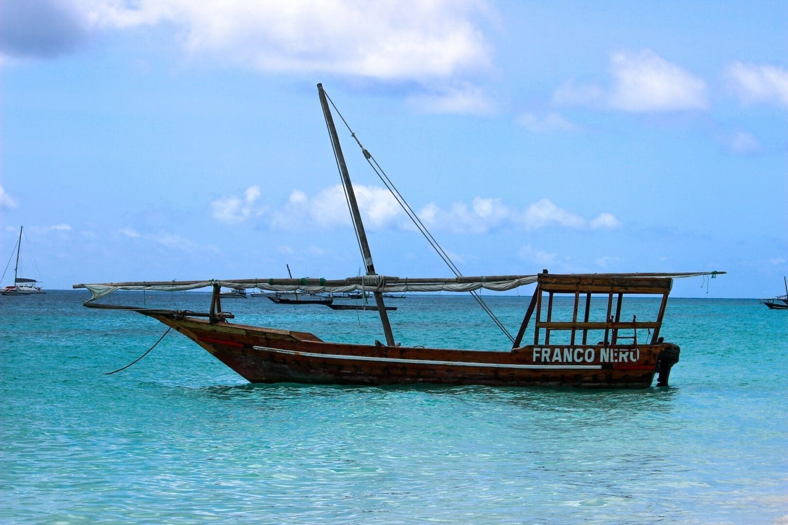 Een houten zeilboot met de naam Franco Nieru dobbert op helder turquoise water voor de kust van Mozambique, met in de verte een paar andere boten zichtbaar onder een gedeeltelijk bewolkte blauwe Afrika-hemel.