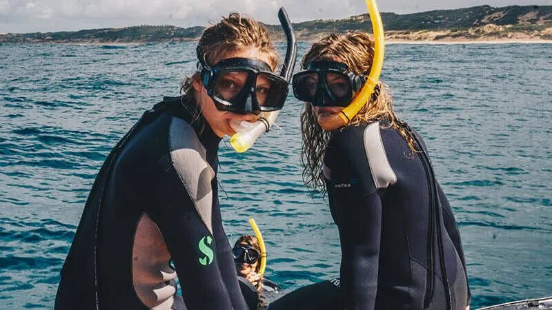 Twee mensen in wetsuits en snorkelspullen zitten op de rand van een boot voor de kust van Mozambique en bereiden zich voor om te gaan snorkelen. De oceaan en de verre kustlijn zijn zichtbaar op de achtergrond onder een gedeeltelijk bewolkte Afrikaanse lucht.
