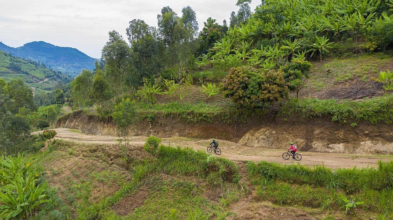Twee fietsers rijden over een kronkelende onverharde weg door weelderige, groene heuvels en landbouwgrond in Rwanda, Afrika, omringd door bomen en bergen in de verte onder een bewolkte hemel.