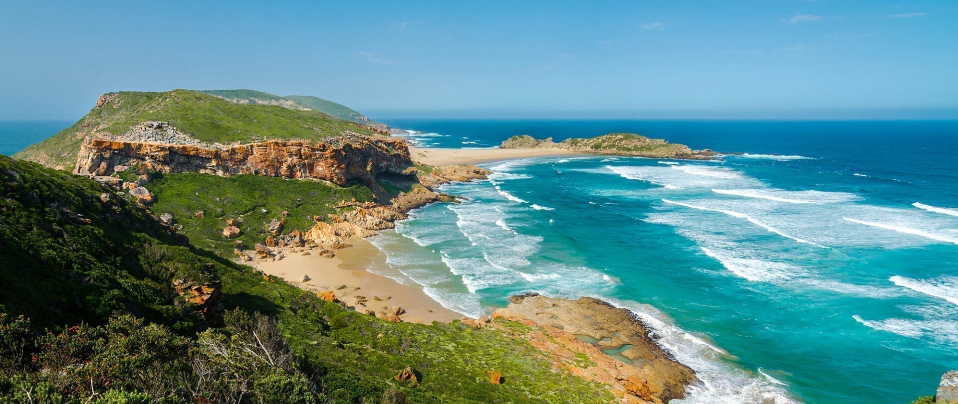 Een schilderachtig kustlandschap in Zuid Afrika met rotsachtige kliffen, groene vegetatie, zandstranden en turquoise golven die onder een strakblauwe hemel langs de kustlijn beuken - een perfecte reisbestemming.