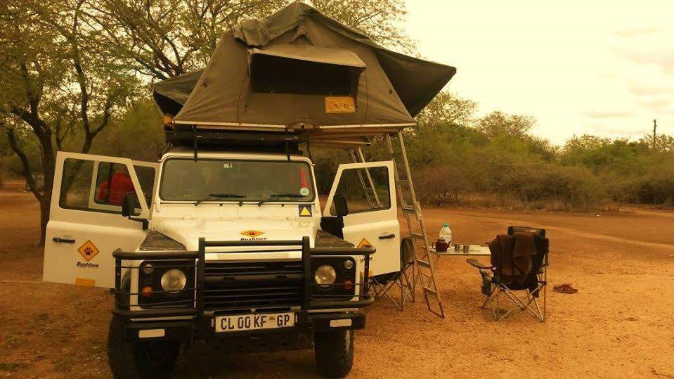 A white off-road vehicle with a roof-top tent is parked at a campsite surrounded by trees in South Africa. Camping chairs, a table with food items, and supplies are set up next to the vehicle, ready for a perfect travelling adventure.