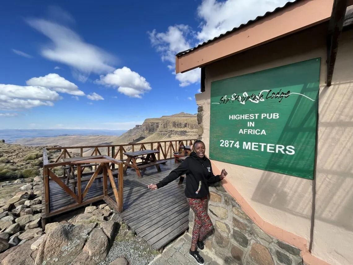 Een glimlachend persoon staat bij een bord met de tekst Highest Pub in Afrika, 2874 meter, bij een houten dek met picknicktafels, met uitzicht op het adembenemende berglandschap van Zuid Afrika onder een blauwe hemel met wolken - perfect voor reisavonturiers.