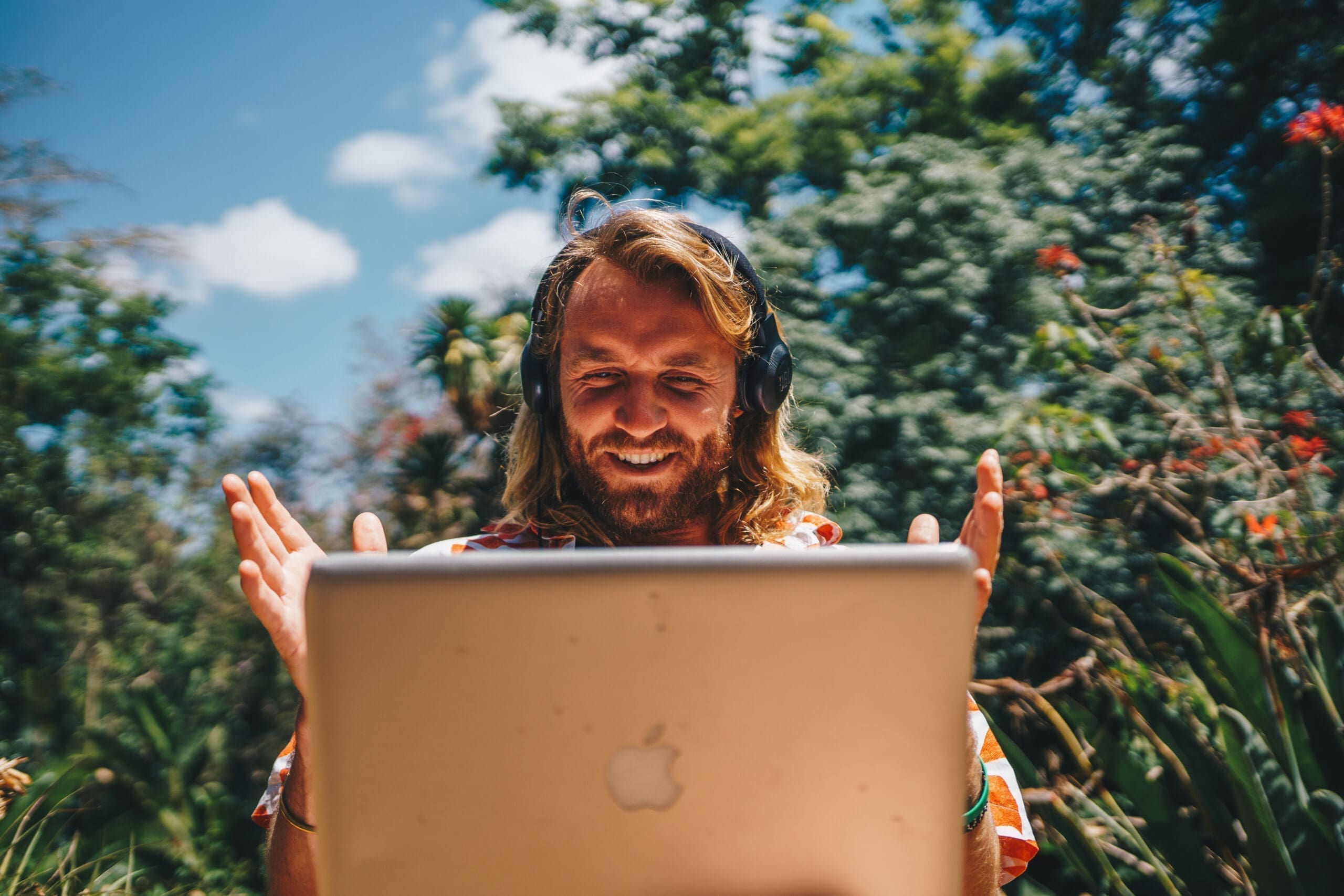 Een man met lang haar en een koptelefoon glimlacht en gebaart naar zijn laptop terwijl hij buiten zit, omringd door weelderig groen en bomen onder een helderblauwe hemel.