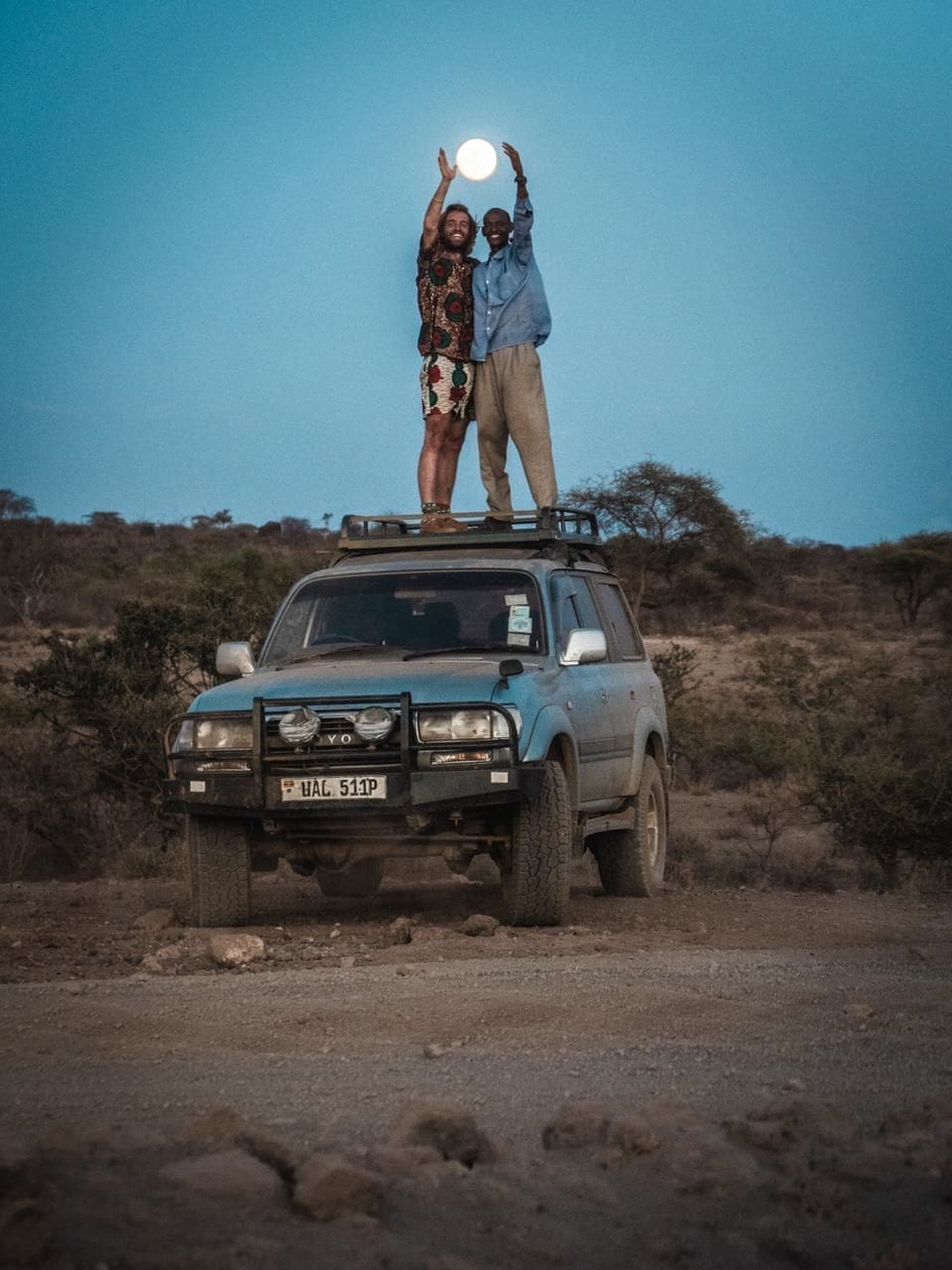 Twee reizigers staan bovenop een geparkeerde SUV in een rotsachtig, open Afrikaans landschap in de schemering. Ze houden hun handen omhoog om de volle maan tussen hen in te kaderen. Struiken en bomen staan op de achtergrond en geven de geest van reizen en avontuur weer.