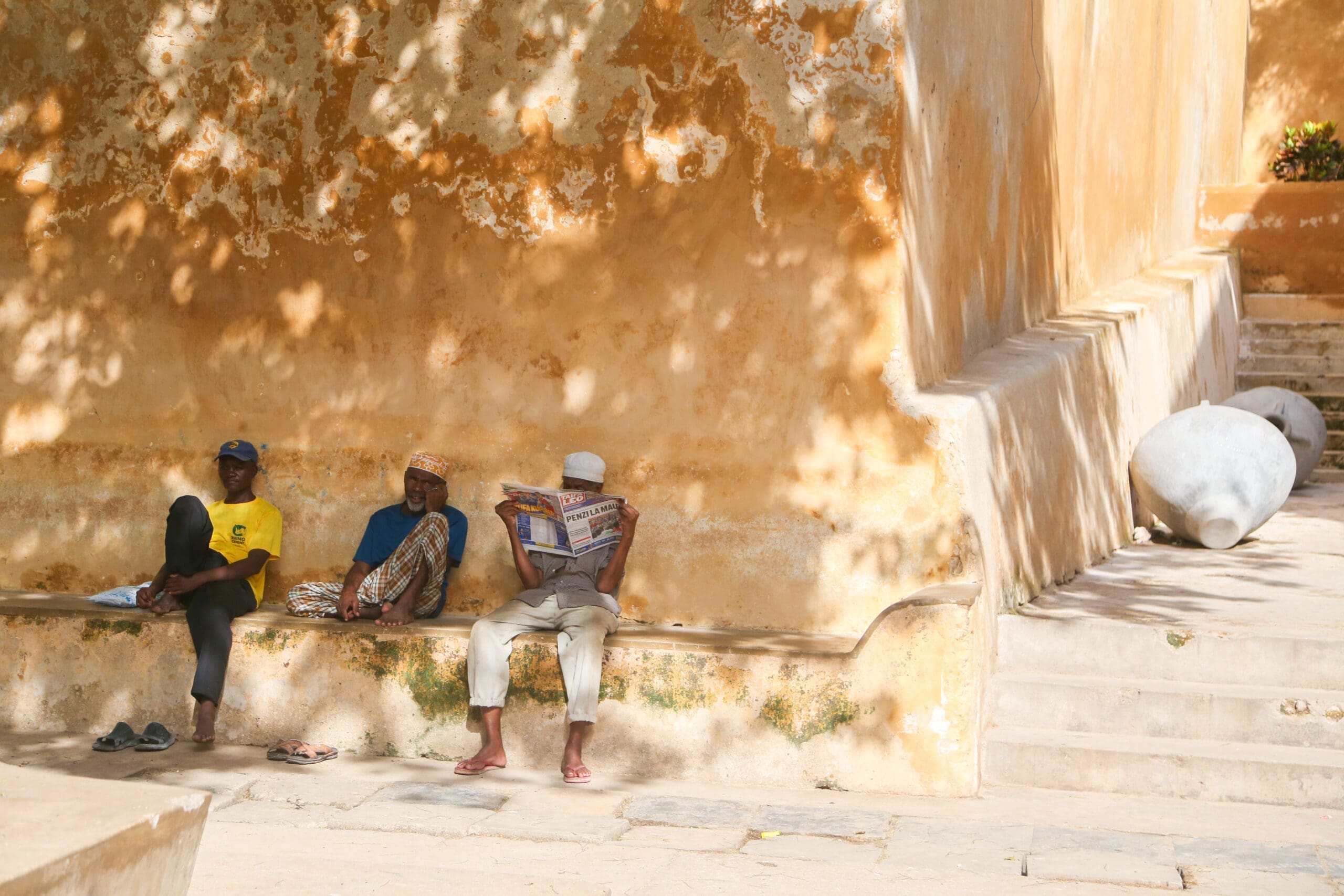 Drie mannen ontspannen tegen een verweerde muur in Lamu terwijl een van hen een krant leest