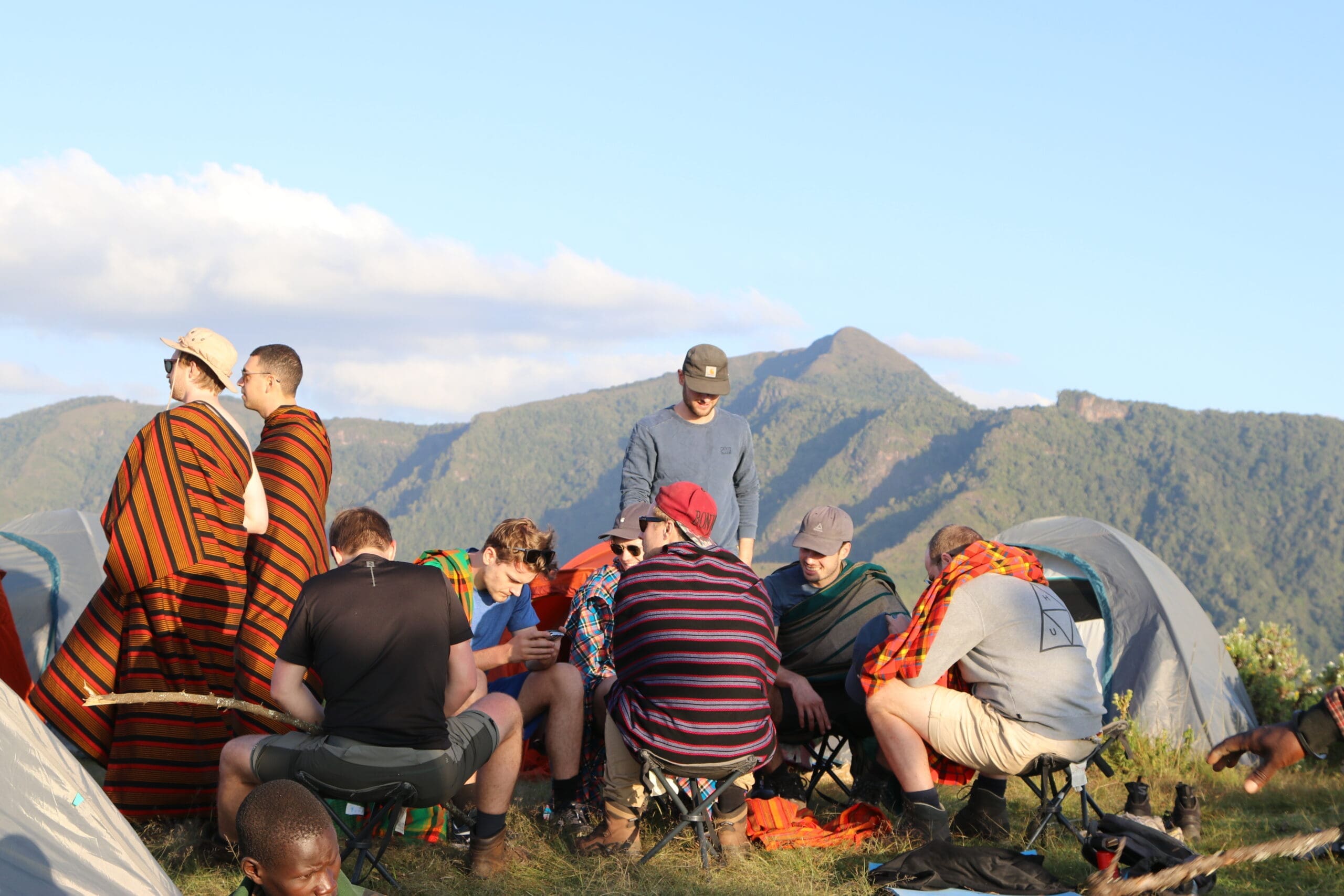 A group of people sit and gather between tents on a grassy hill in Uganda, with mountains in the background. Some wear striped blankets, enjoying the clear, sunny sky during their travelling adventure in Africa.