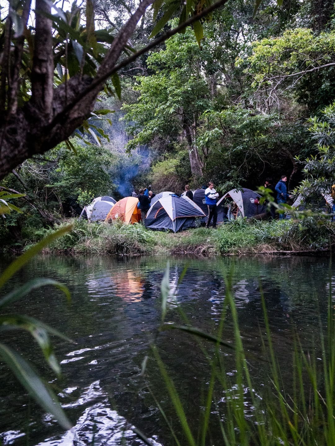 een groep mensen kampeert met tenten aan de rand van een beboste rivier in eswatini terwijl rook opstijgt van een kampvuur tussen de bomen