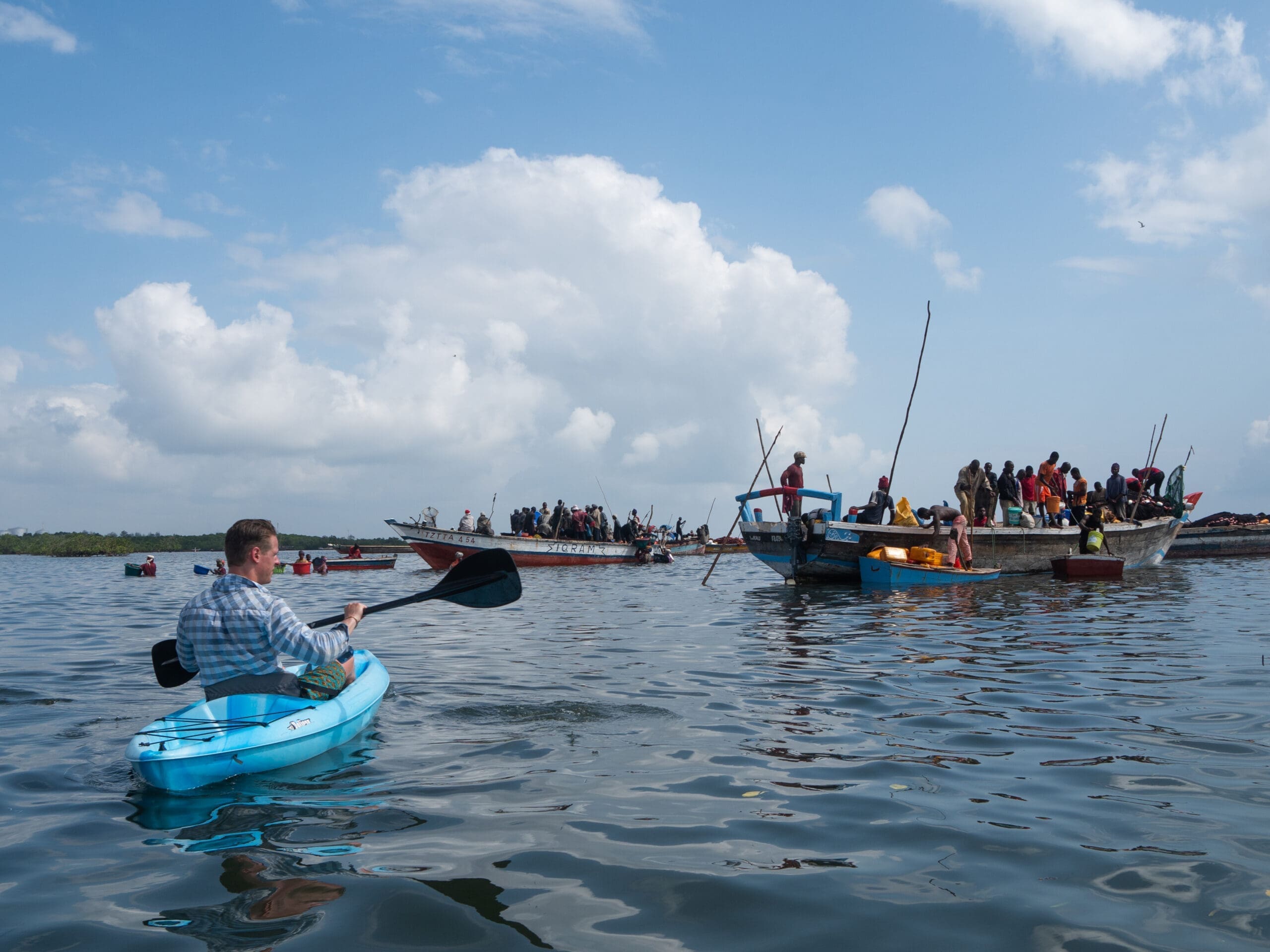 Een persoon in een blauwe kajak peddelt over kalm water in de richting van een aantal vissersboten vol met mensen onder een gedeeltelijk bewolkte hemel, die de serene geest van reizen in Afrika vastlegt.