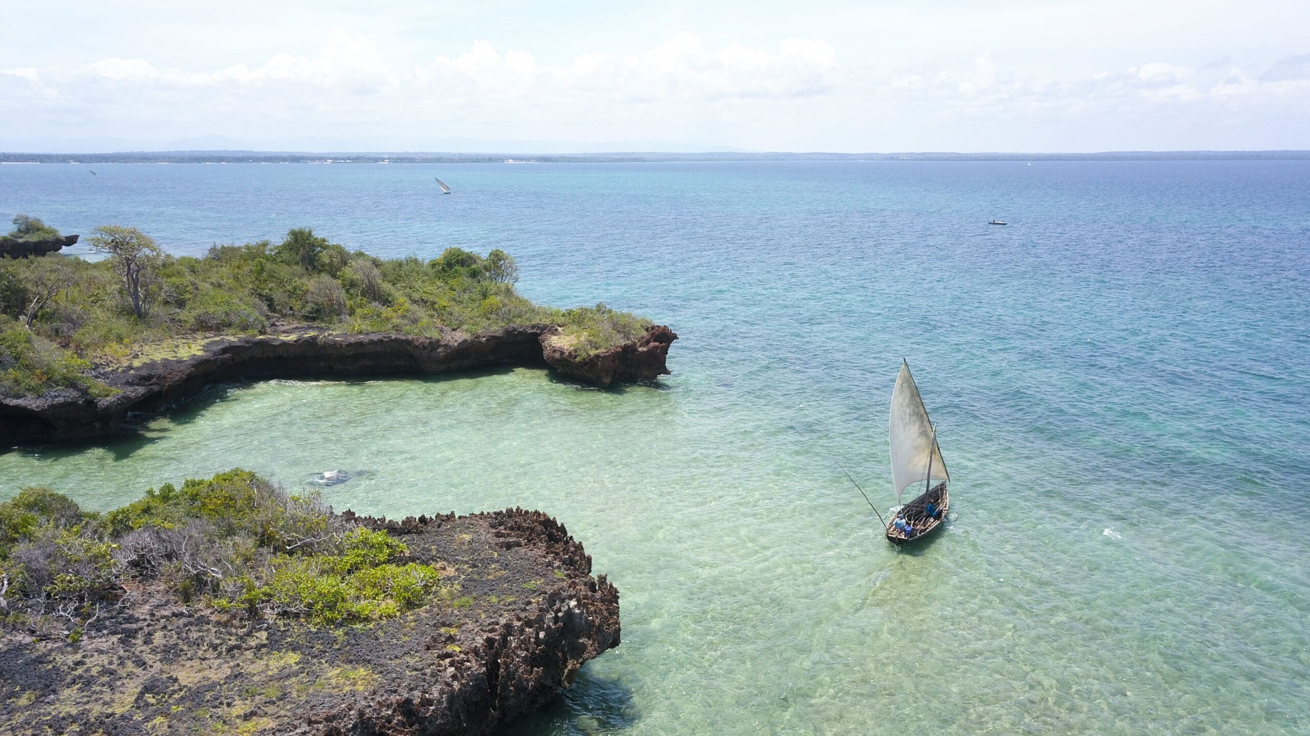 Een klein zeilbootje met een enkel wit zeil glijdt over helder turquoise water in de buurt van de rotsachtige, groene kustlijn van Tanzania onder een heldere, halfbewolkte hemel - een perfect tafereel voor wie houdt van reizen in Afrika.