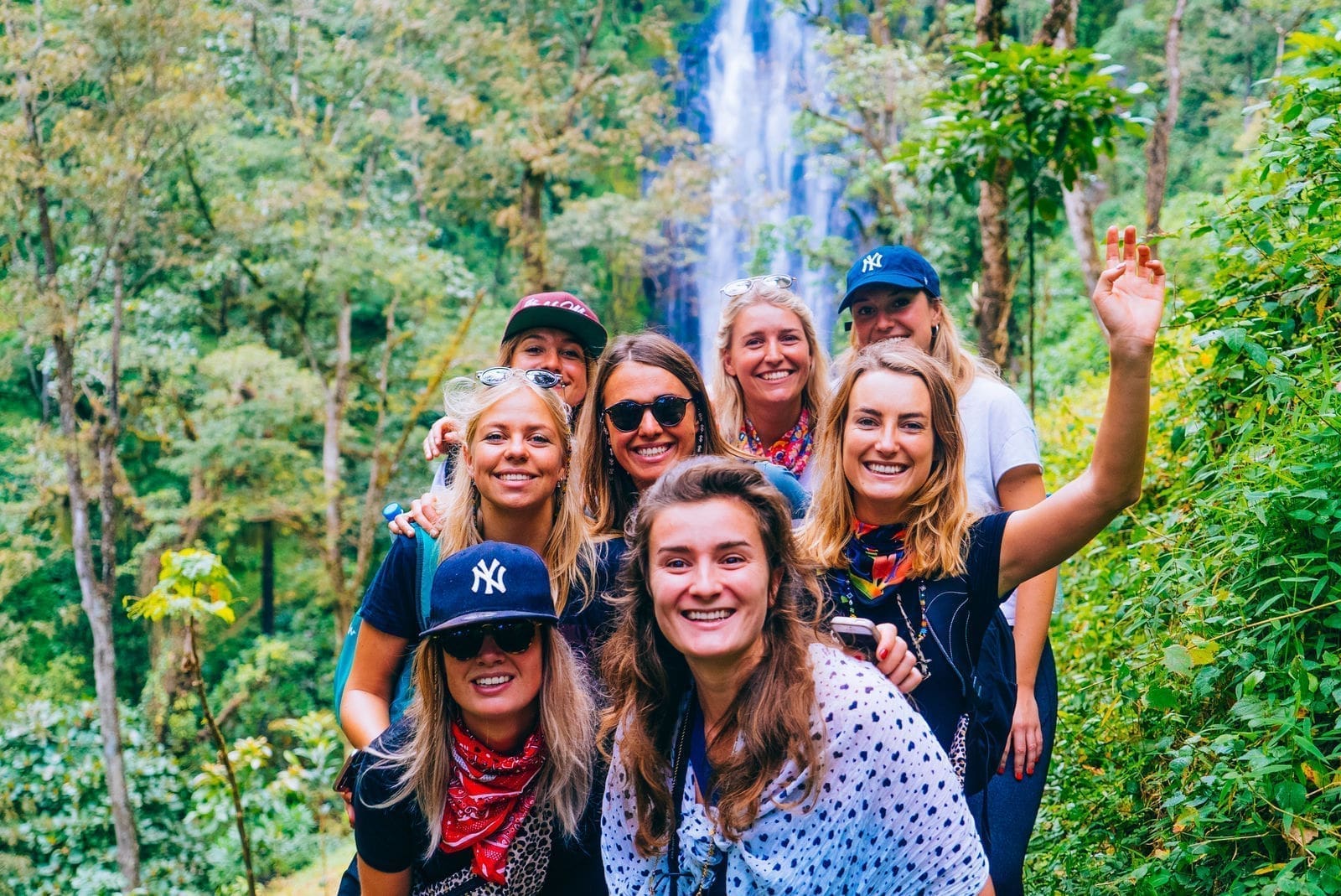 Een groep lachende jonge vrouwen poseert samen op een groen bospad met een hoge waterval op de achtergrond. Sommigen dragen zonnebrillen, petjes en rode bandana's, en eentje zwaait naar de camera.