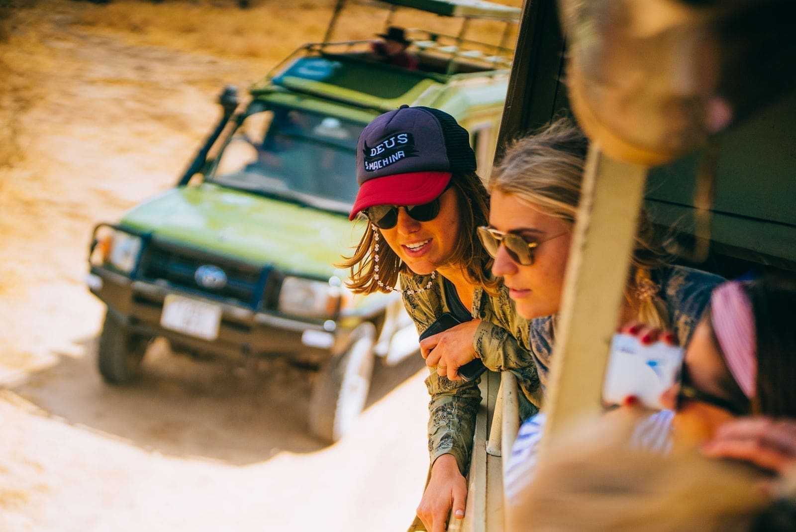 Drie reizigers leunen uit een safarivoertuig, glimlachen en kijken naar iets buiten. Een andere groene safari-jeep is zichtbaar op de achtergrond op een onverharde weg in zonnig Tanzania.