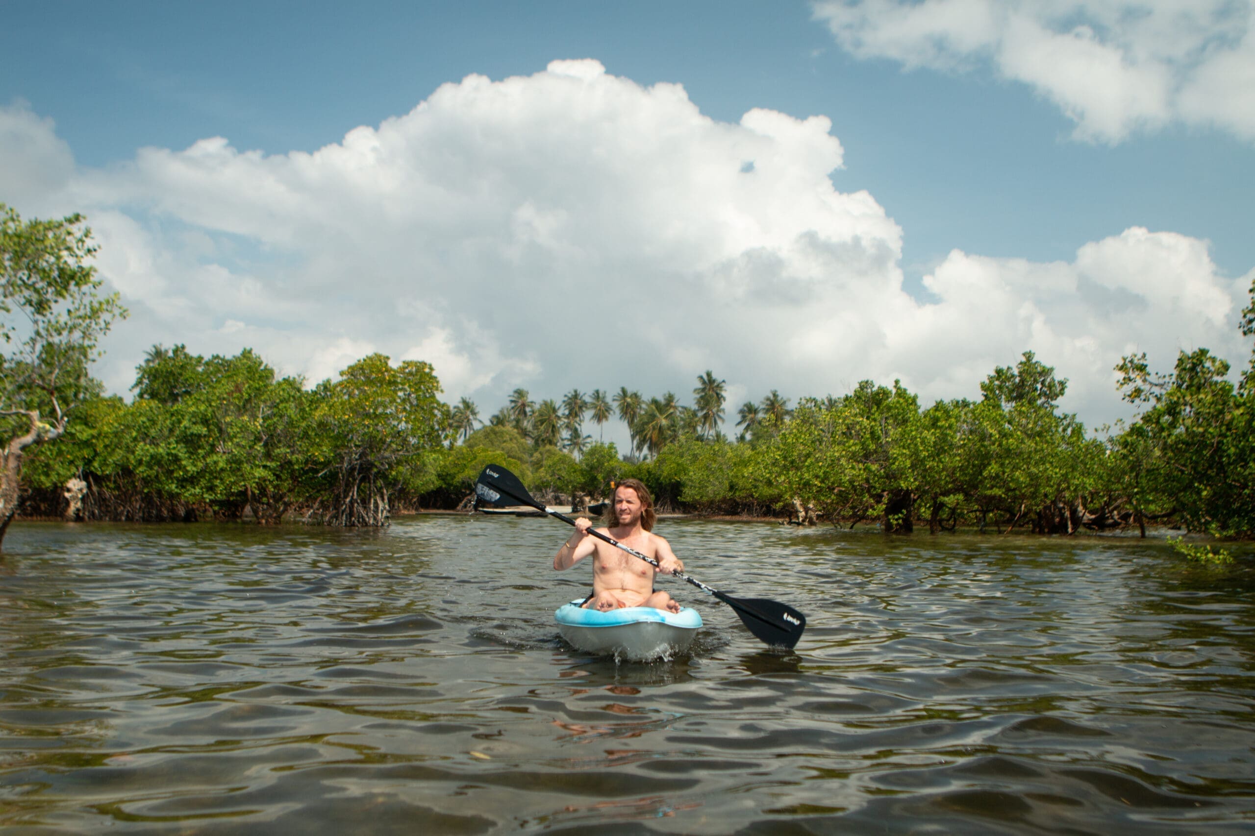Een persoon peddelt in een kajak op kalm water omringd door groene mangroves onder een blauwe lucht met grote, pluizige wolken in Tanzania. Op de achtergrond zijn palmbomen te zien, wat zorgt voor een ideaal Afrika reizen avontuur.