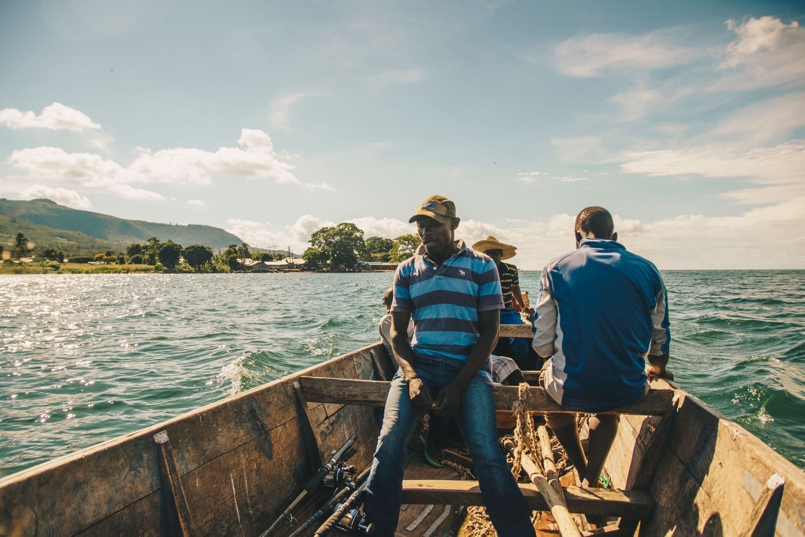 Drie mannen zitten in een houten boot op een blauw meer, met zonlicht dat weerkaatst op het water en groene heuvels zichtbaar op de achtergrond onder een gedeeltelijk bewolkte hemel.