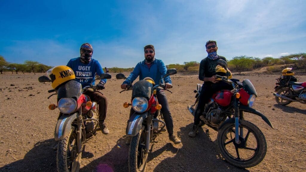 Drie mannen met zonnebrillen en bandana's zitten op motoren in een droog, open landschap met weinig bomen en een strakblauwe lucht. Rechts op de achtergrond staan nog twee motorfietsen.