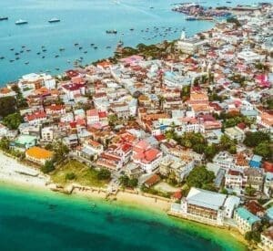 Luchtfoto van een kuststad in Tanzania met kleurrijke daken, een wit zandstrand, turquoise water en veel boten die voor de kust voor anker liggen. Het stadsbeeld wordt gekenmerkt door drukke gebouwen en weelderig groen langs de kustlijn - perfect voor reizen in Afrika.