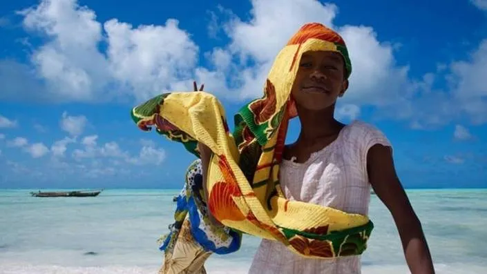 Een lachend kind in een witte jurk staat op een strand in Tanzania en houdt een kleurrijke stof in de wind. De oceaan en een kleine boot zijn zichtbaar op de achtergrond onder een helderblauwe lucht met verspreide wolken - een perfect reismoment.