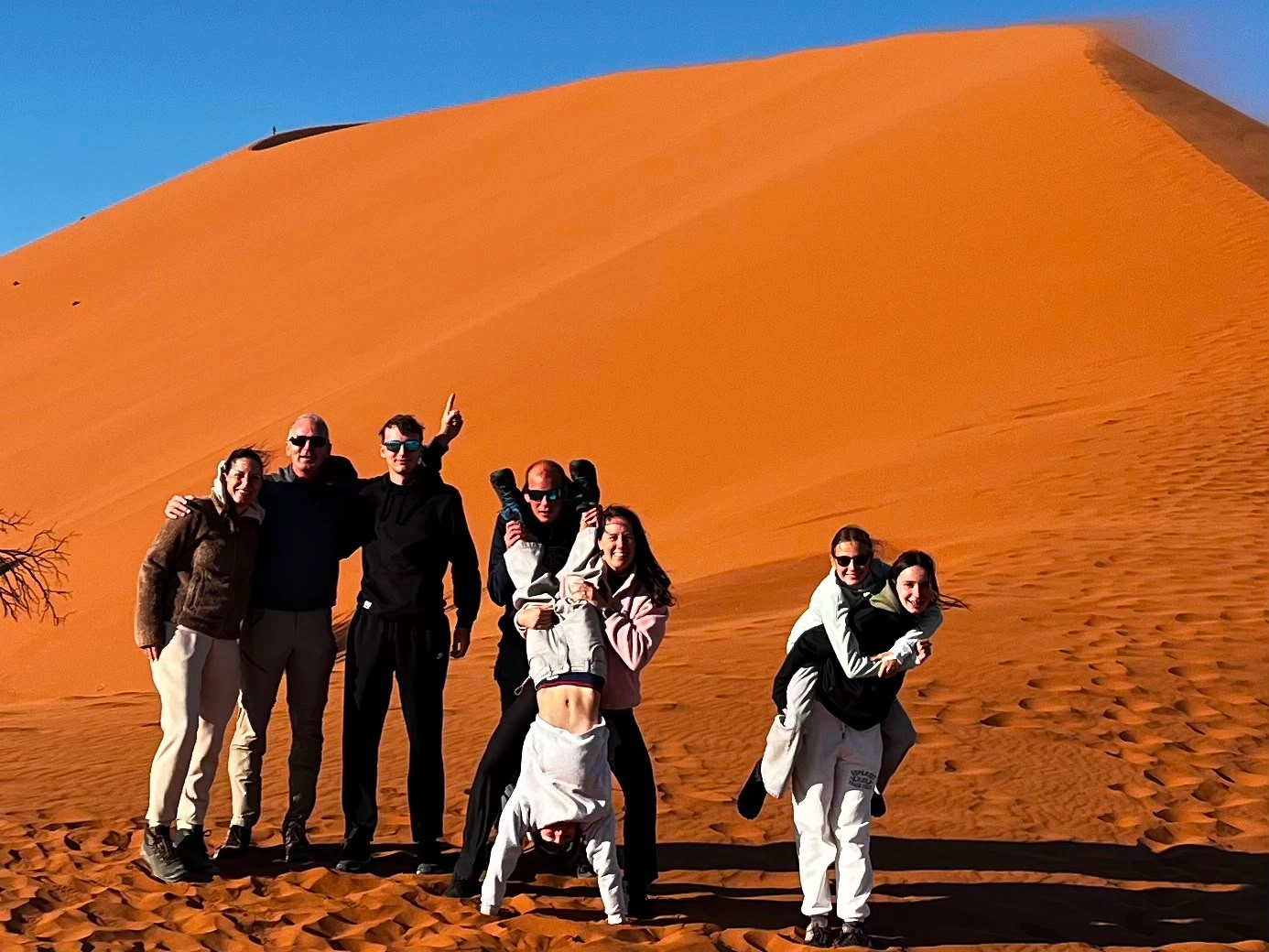 Een groep van acht mensen poseert speels op oranje zandduinen onder een strakblauwe hemel; één persoon doet een handstand, twee anderen dragen iemand op hun rug en de rest staat dicht bij elkaar en glimlacht.