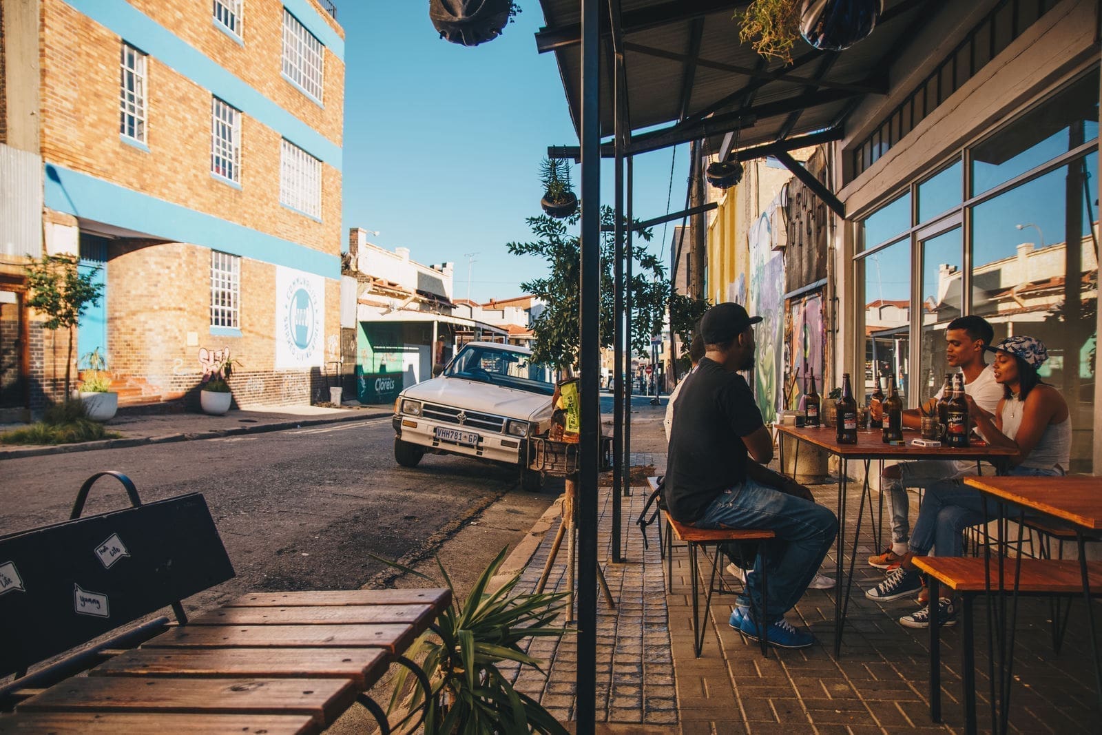 Mensen zitten aan een cafétafel op een stoep in de stad te drinken en te praten. Vlakbij staat een oldtimer geparkeerd en het tafereel speelt zich af tegen bakstenen gebouwen onder een strakblauwe hemel.