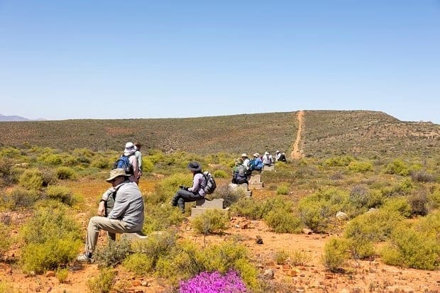 Mehrere Wanderer mit Hüten und Rucksäcken sitzen in Abständen auf Felsen oder Bänken entlang eines Weges in einer trockenen, mit Sträuchern bewachsenen Landschaft unter einem klaren blauen Himmel.