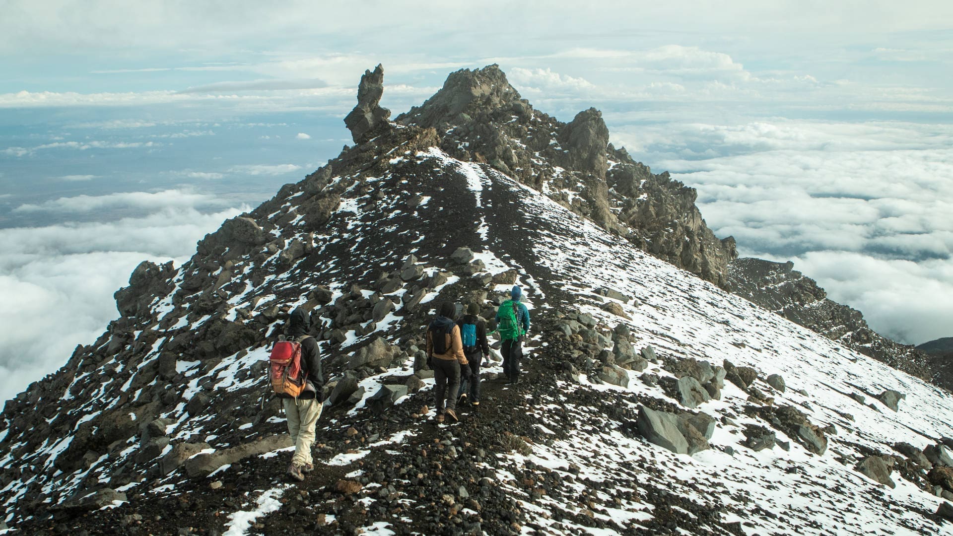 Vier wandelaars met rugzakken lopen over een rotsachtige, besneeuwde bergrug boven de wolken in Tanzania, op weg naar grillige pieken onder een gedeeltelijk bewolkte hemel - een onvergetelijke Afrika reizen ervaring.