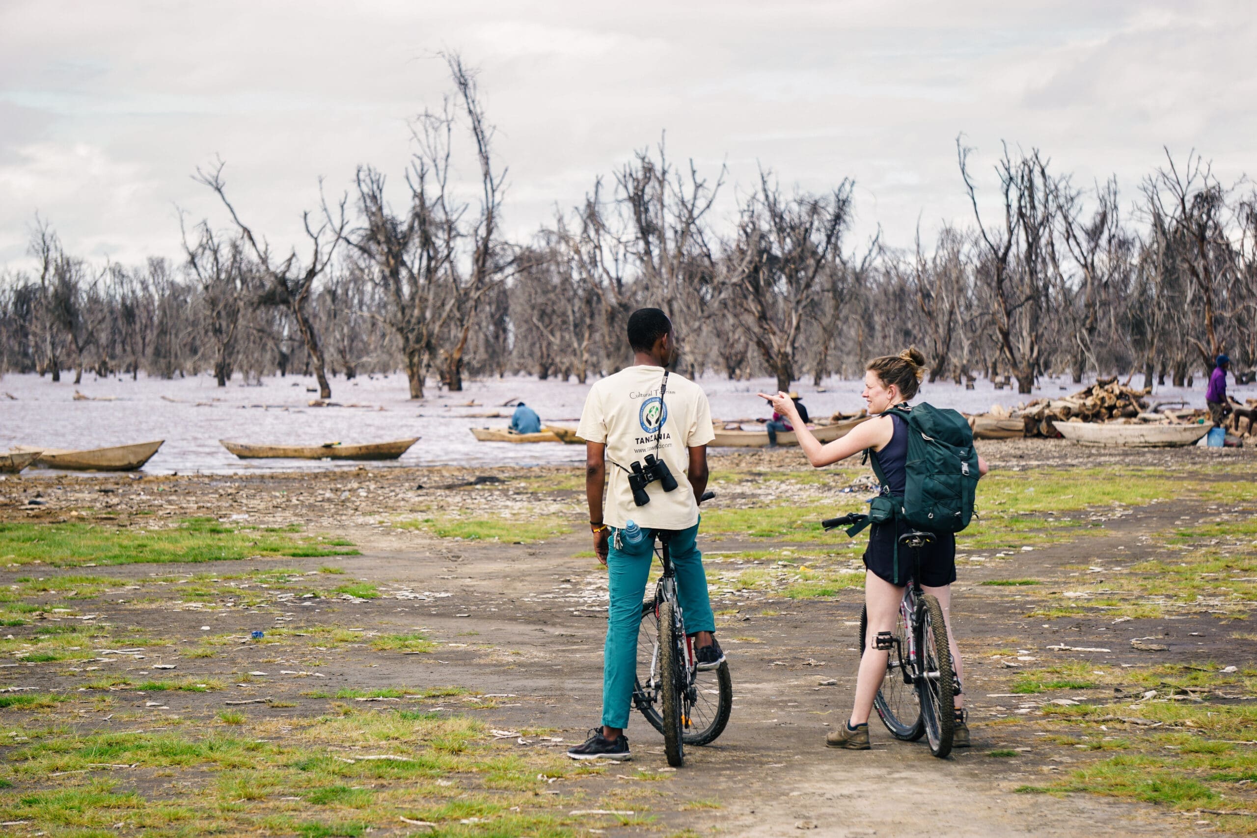 Twee fietsers stoppen bij een meer met dorre, bladerloze bomen en houten boten op de achtergrond; de een gebaart terwijl ze praten, beiden dragen rugzakken en camera's.