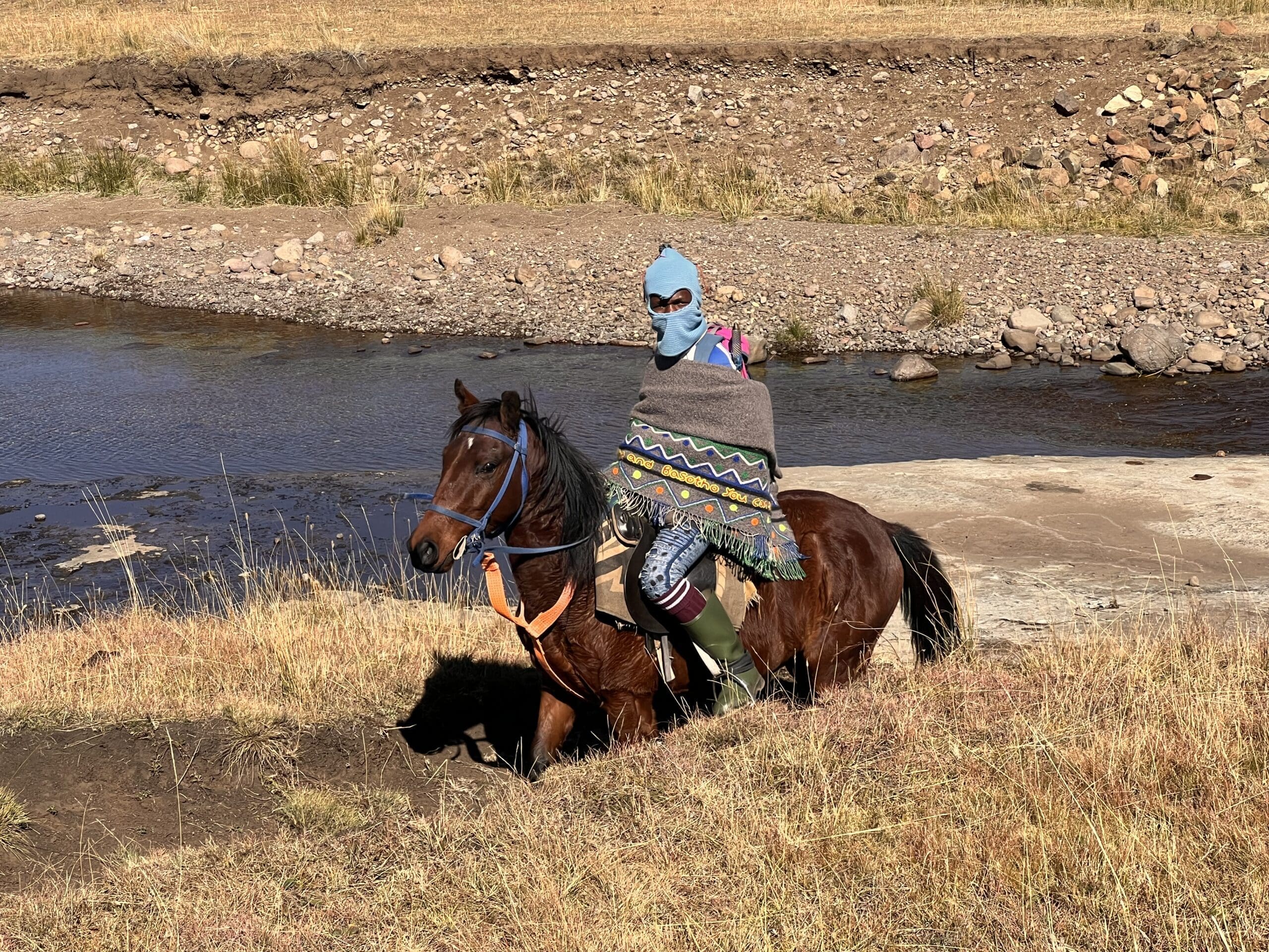 Een persoon met een blauwe bivakmuts, poncho met patroon en laarzen rijdt op een zonnige dag op een bruin paard door droog gras bij een ondiepe rivier.