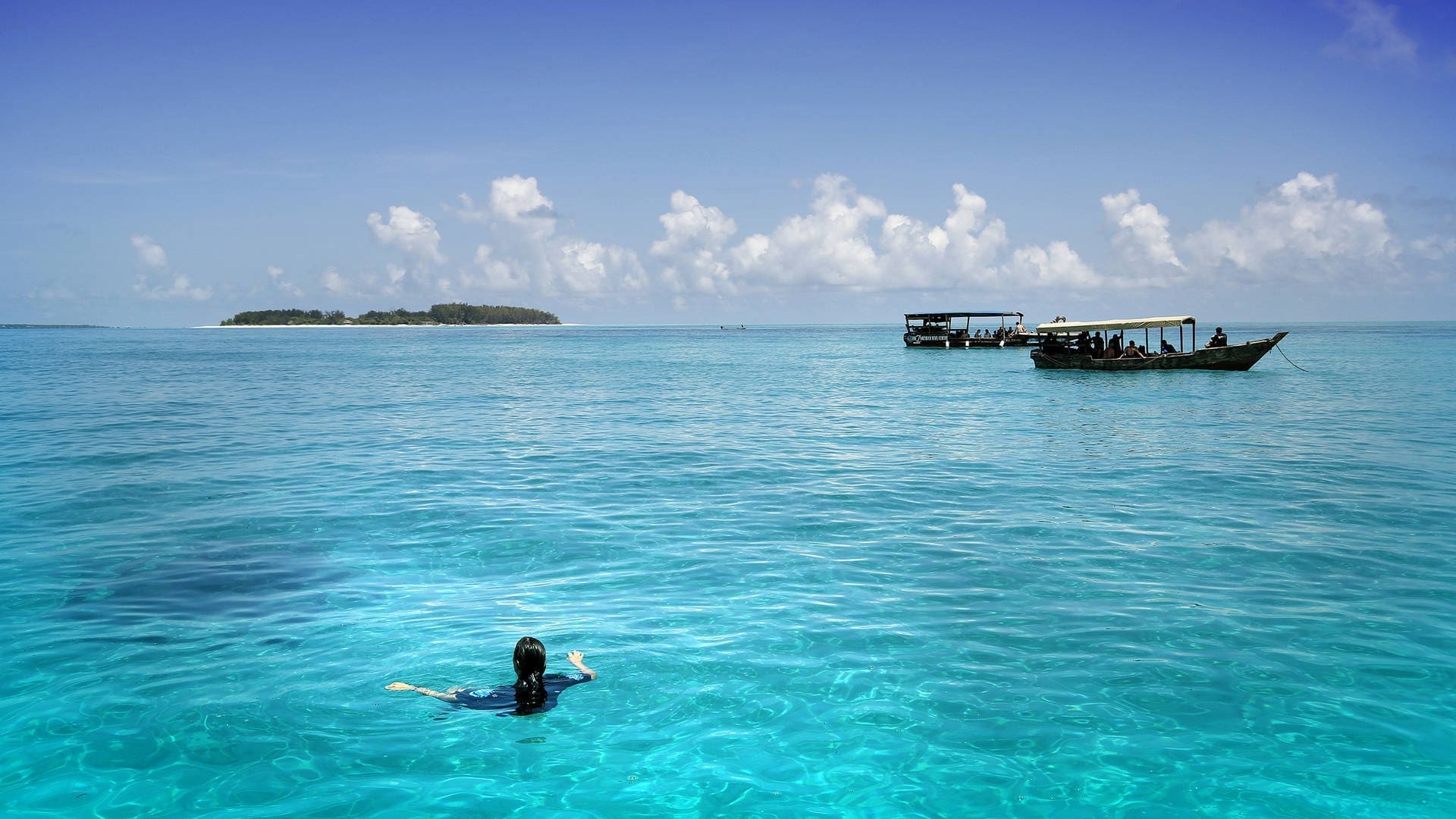 Een persoon zwemt in helderblauw oceaanwater bij een houten boot en hut in de verte, met een eiland aan de horizon onder een heldere hemel - een perfect reizen tafereel in Tanzania, Afrika.