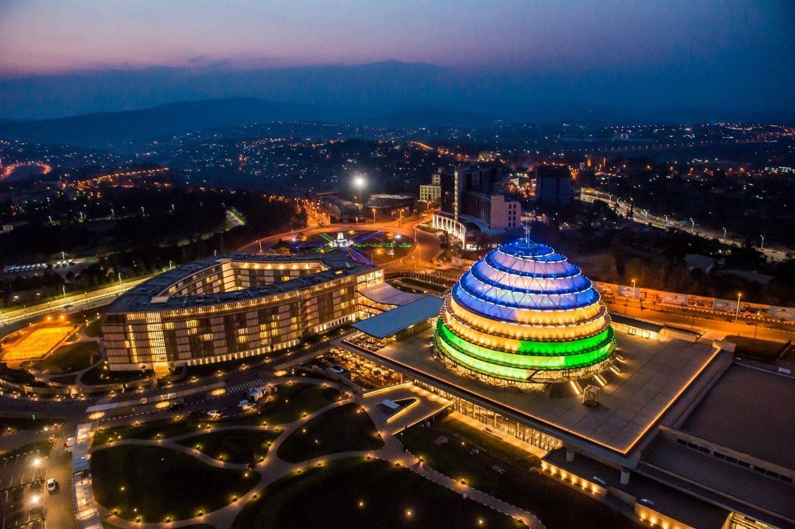 Luchtfoto van een modern koepelvormig congrescentrum in Rwanda, verlicht met blauwe, groene en gele lichten, omringd door stadsgebouwen in de schemering - een levendig herkenningspunt in het hart van Afrika.