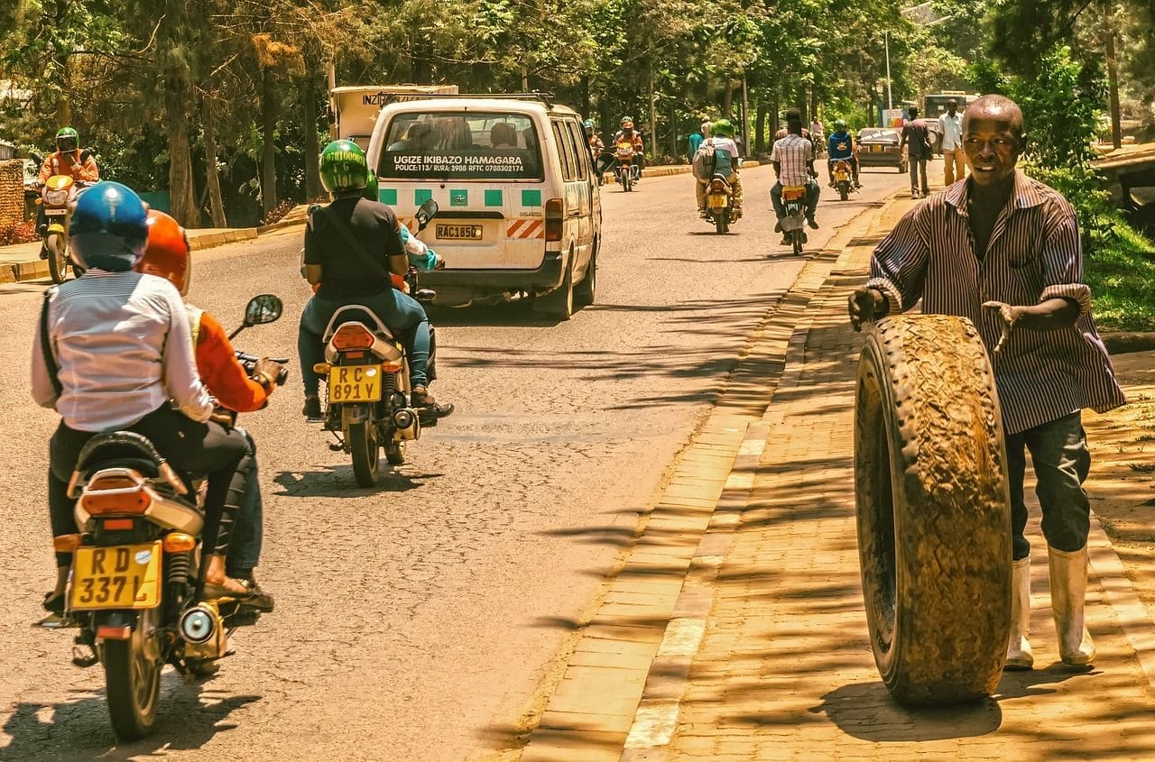 Een man rolt een grote, versleten band op een trottoir in Rwanda terwijl motoren en een busje op een zonnige dag door een drukke, met bomen omzoomde straat rijden.