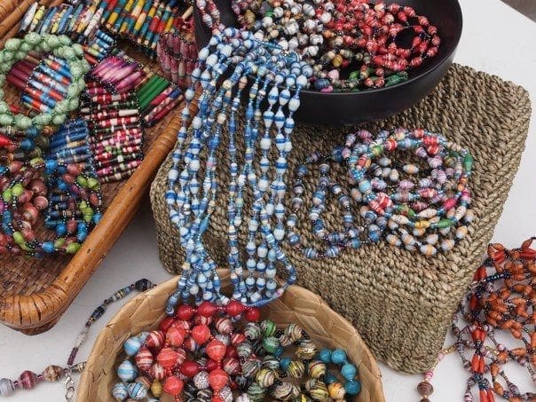 Kleurrijke handgemaakte kralenkettingen en armbanden uit Oeganda, sommige in schalen en manden, uitgestald op geweven dienbladen. Perfect voor reizigers die houden van levendige patronen en unieke souvenirs van hun reizen.