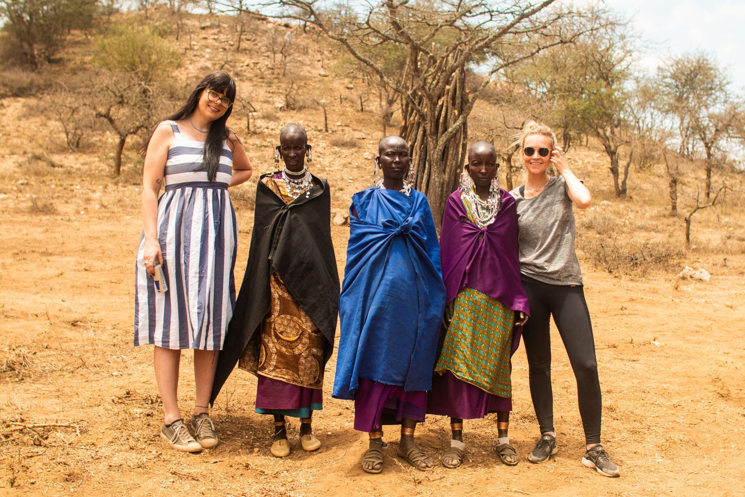 Two women in casual clothes stand with three Maasai women in traditional colorful clothing and jewelry, posing together outdoors in a dry, hilly Tanzania landscape—a memorable travelling experience in Africa.