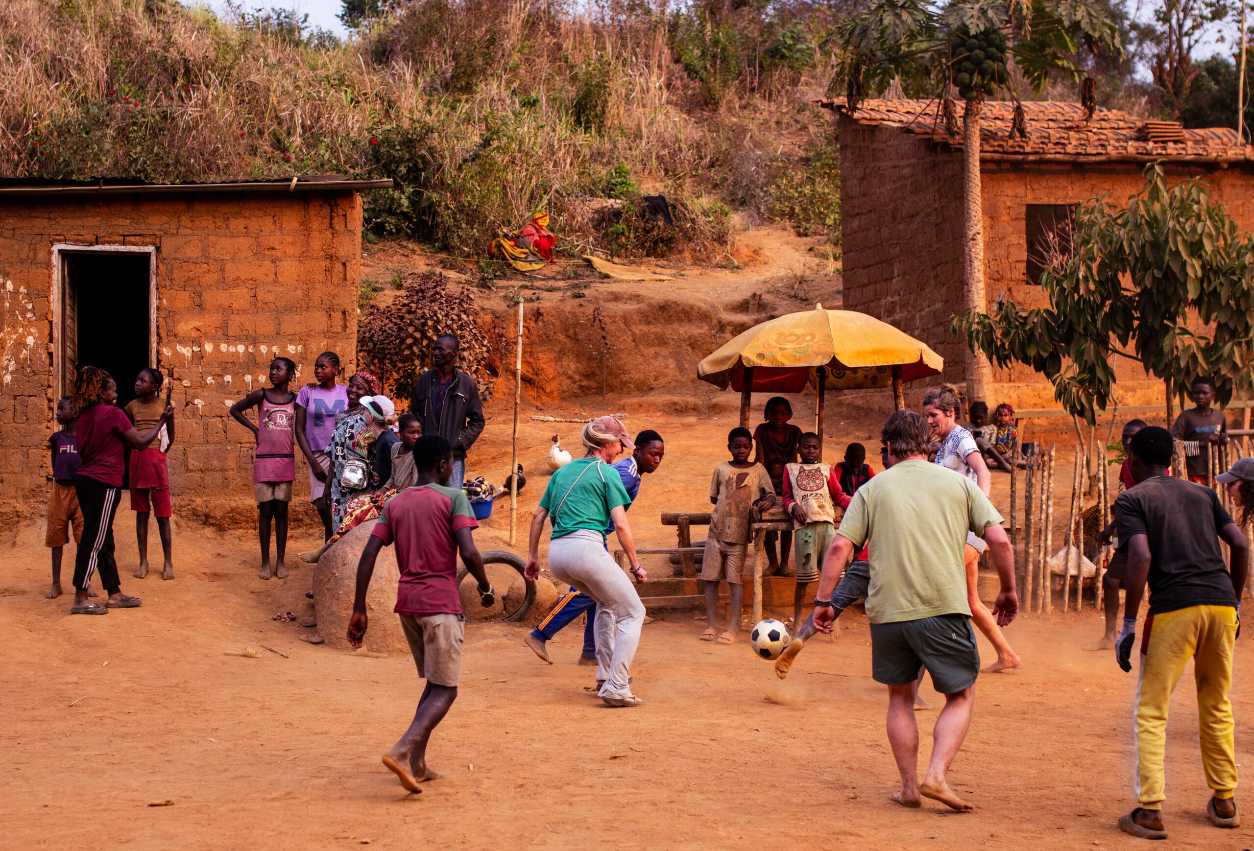 Een groep kinderen en volwassenen speelt voetbal op een zandveld in een plattelandsdorp in Angola, omringd door eenvoudige stenen huizen en bomen, terwijl reizigers toekijken en socialiseren op de achtergrond.
