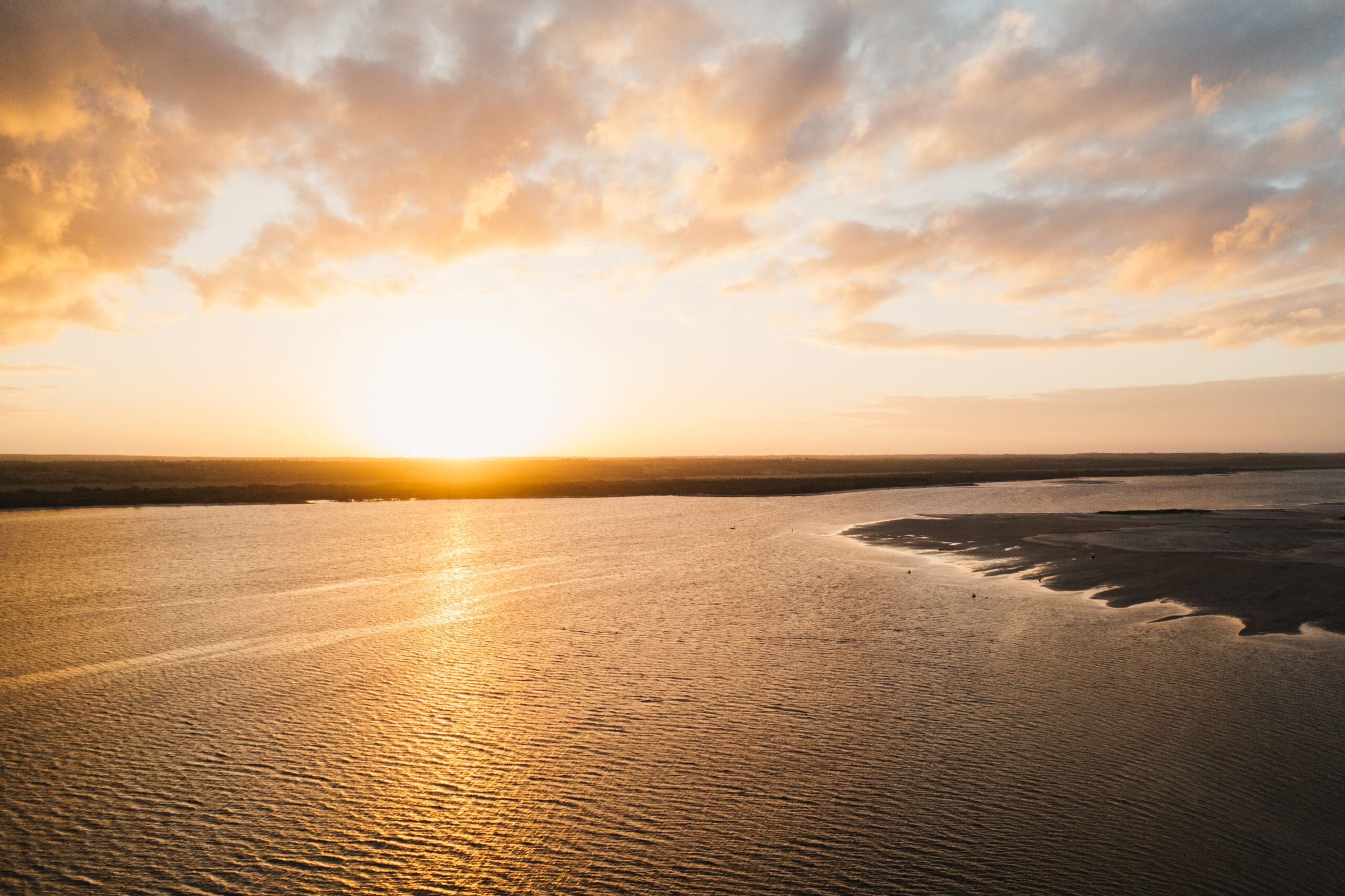 Een serene zonsondergang over een brede rivier of meer in Mozambique, Afrika, met gouden zonlicht dat reflecteert op het water en een zanderige kustlijn aan de rechterkant. Zachte wolken vullen de lucht en creëren een vredige en warme sfeer.