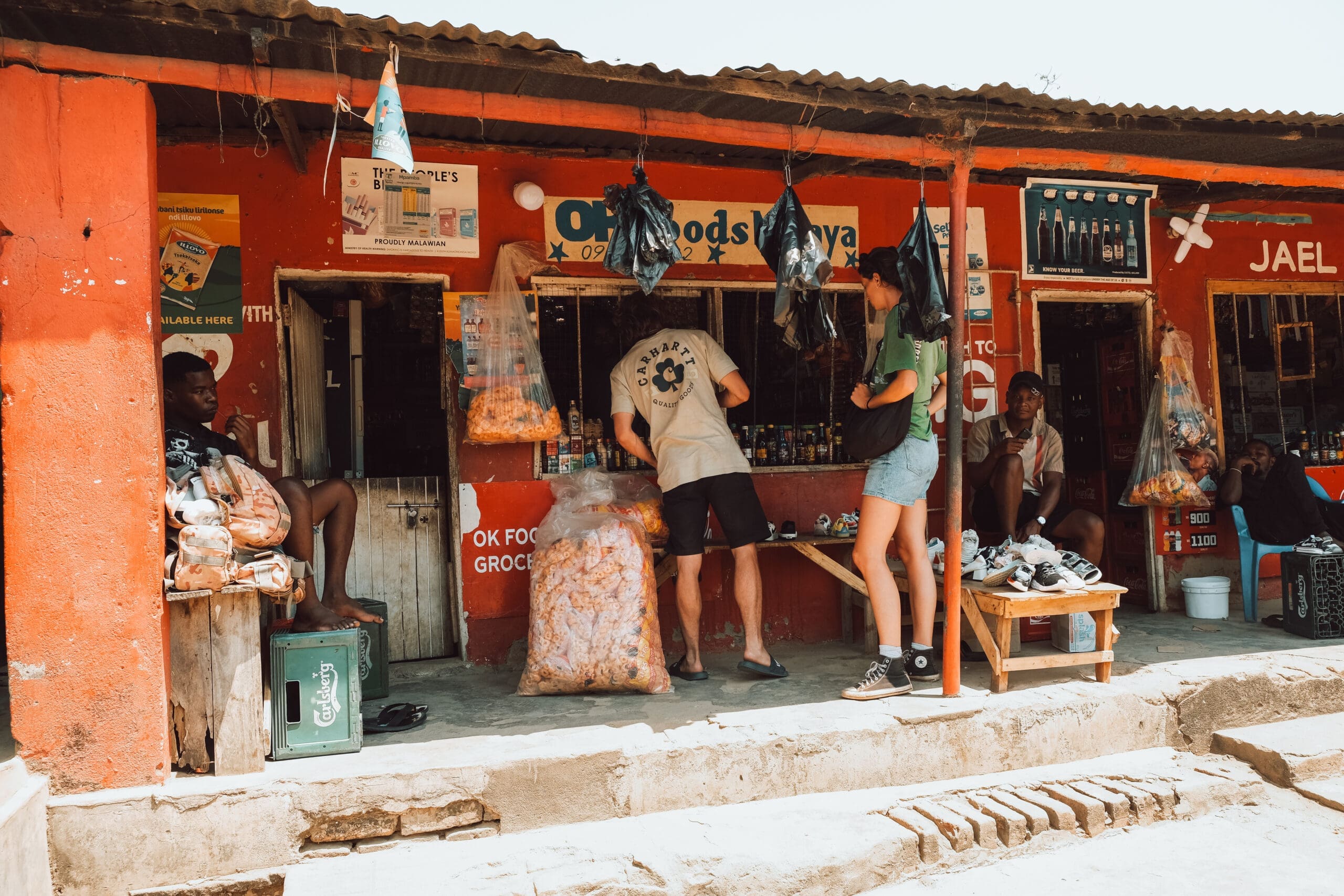 A small, red-painted grocery shop in Malawi with snacks hanging outside. People stand and interact near the counter; some are seated nearby. The lively scene, brightened by sunlight, captures the spirit of travelling through Africa.