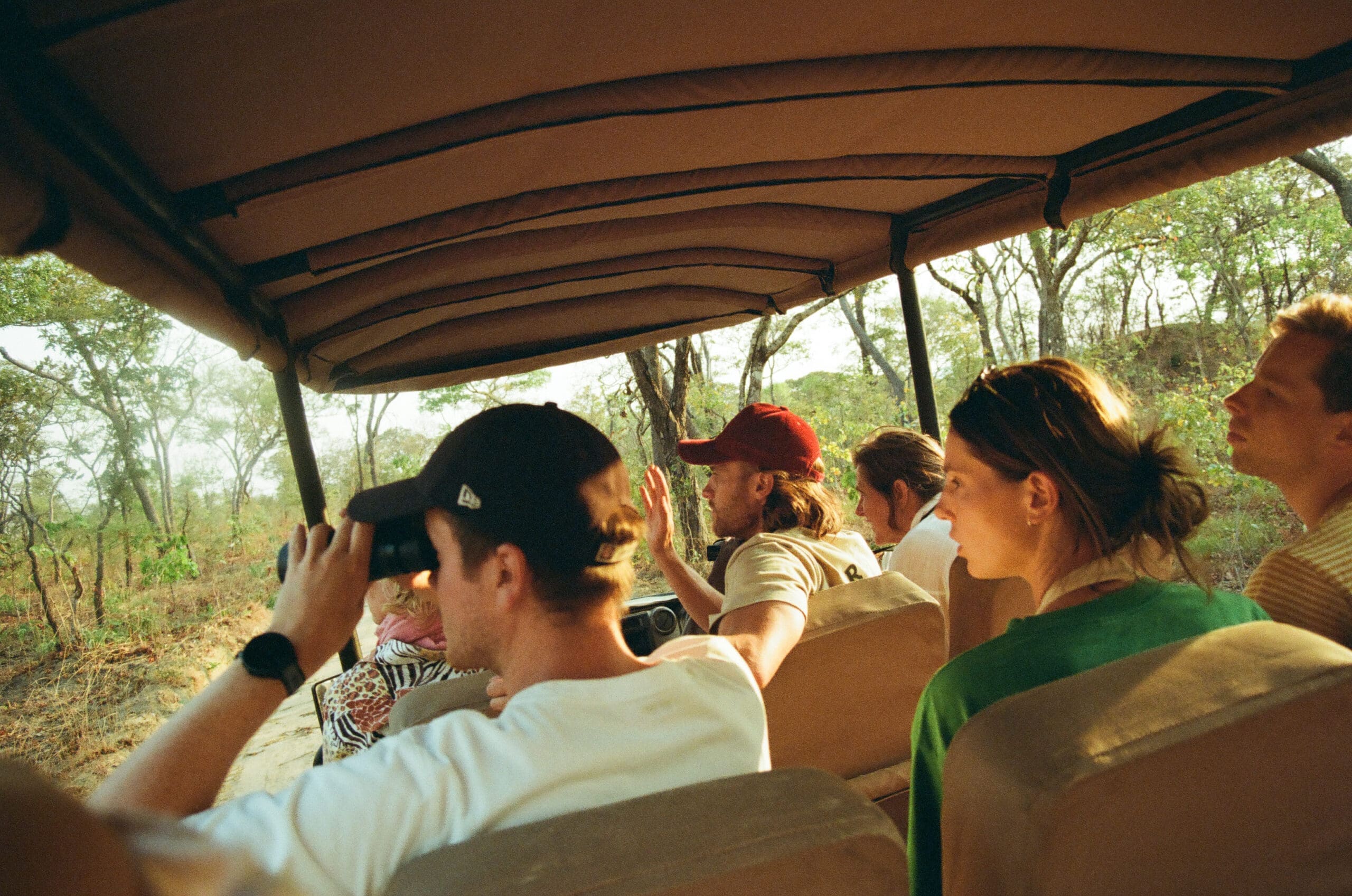 Een groep mensen zit in een open safarivoertuig en verkent de bosrijke schoonheid van Malawi. Eén persoon gebruikt een verrekijker, terwijl anderen het landschap bewonderen. Zonlicht filtert door de bomen en legt de magie van Afrika reizen vast.