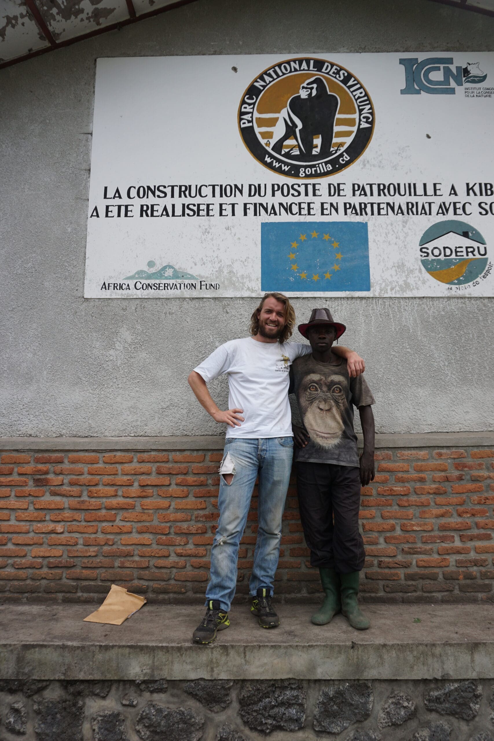 Twee mannen staan lachend naast elkaar voor een muur met een bord van het Virunga National Park en verschillende natuurbeschermingslogo's. De ene man draagt een wit shirt en spijkerbroek, de andere een shirt met gorillaprint, een hoed en groene laarzen. De ene man draagt een wit shirt en een spijkerbroek, de andere een shirt met gorillaprint, een hoed en groene laarzen.