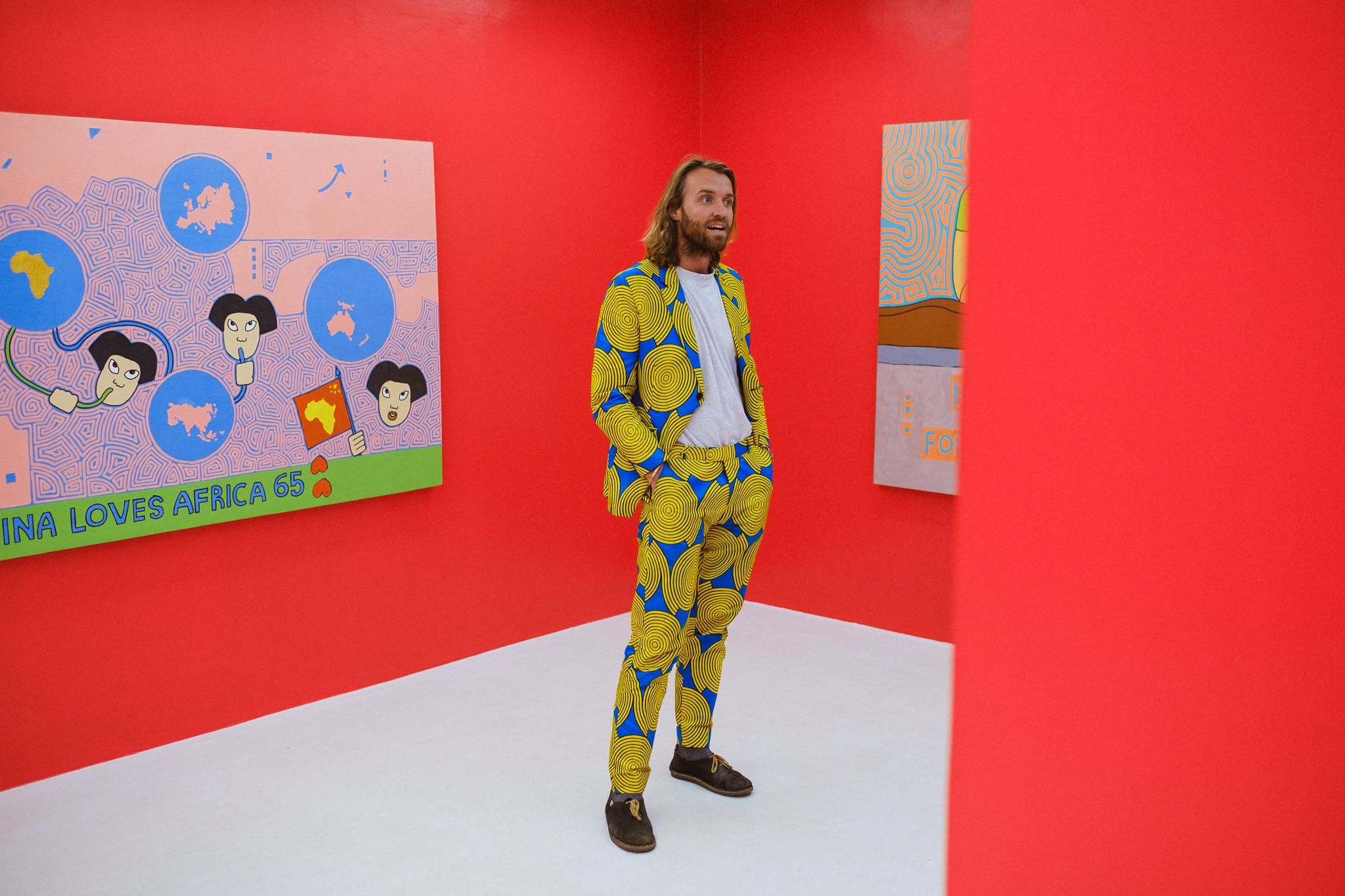 Een man met lang haar en een baard staat in een felverlichte galerie met rode muren en draagt een kleurrijk pak met gele en blauwe patronen. Kunstwerken met abstracte gezichten en landkaarten hangen aan de muren om hem heen.