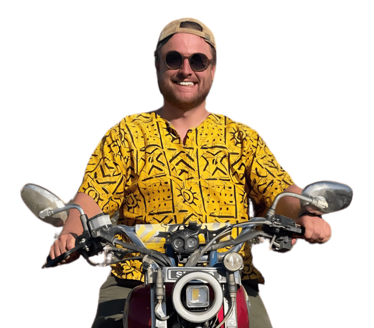 Een lachende man met zonnebril, beige pet en felgeel overhemd met patroon zit op een motorfiets, houdt het stuur vast, met een witte achtergrond.